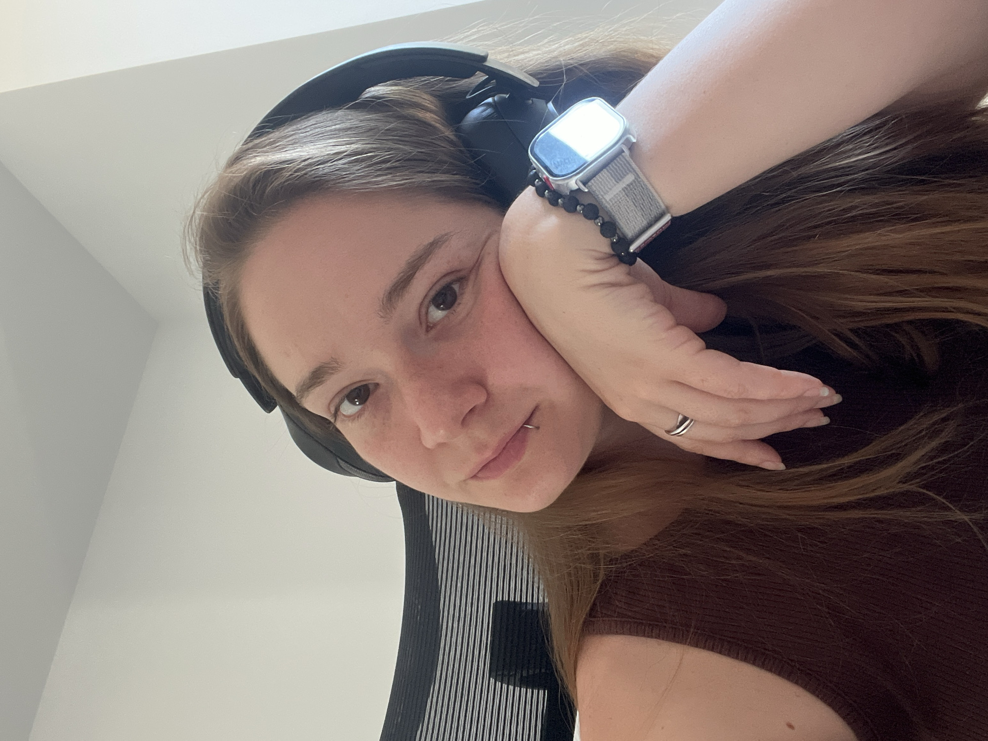 A young woman with light brown hair and freckles is sitting in an office chair, wearing headphones and resting her head on her hand. She has a piercing in her lower lip and is wearing a brown tank top, a ring on her finger, and a smartwatch with a grey band. The background is a plain white wall, and a black mesh office chair is visible behind her. There is no indication of the city of Plovdiv, Bulgaria in the image.