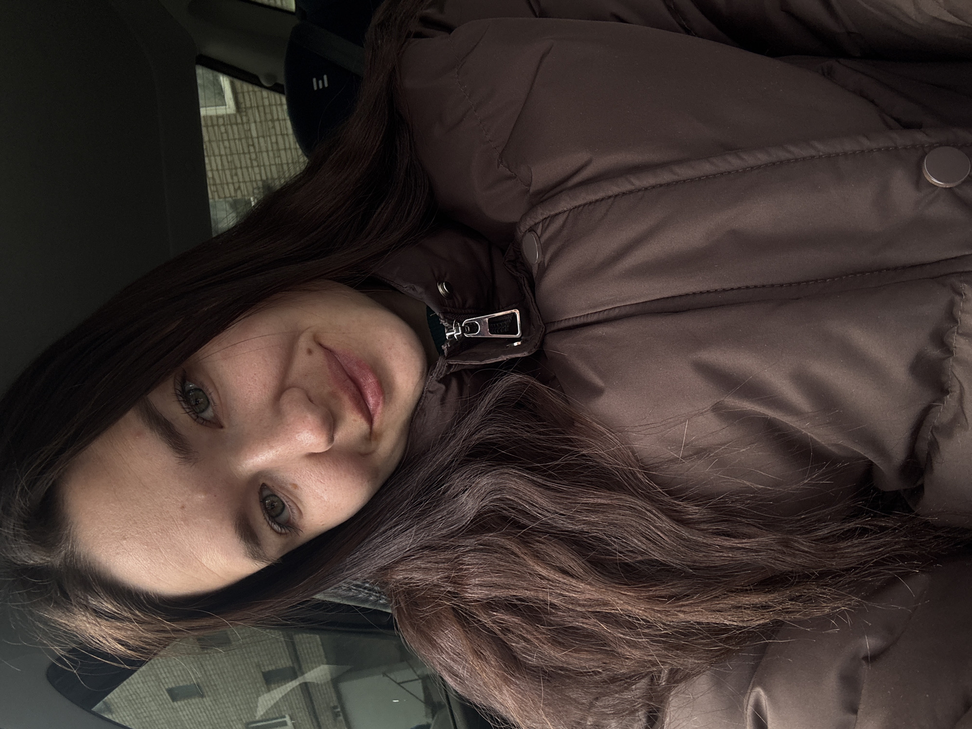 A young woman with long, dark brown hair and light-colored eyes is wearing a brown puffer jacket. She is inside a vehicle, possibly a car, as the interior is visible. Outside the window, a building with brickwork can be seen. Based on the metadata, this image was taken in the city of Chaikovsky, Russia.