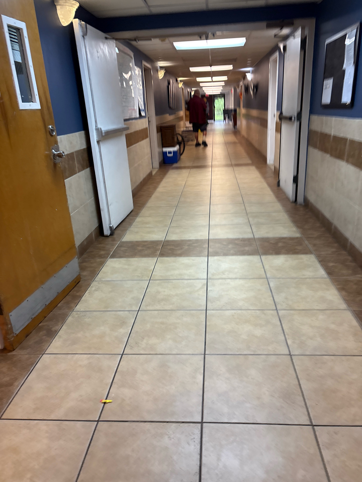 The image shows a long, tiled hallway in a building located in South Bradenton, United States of America. The hallway has doors on both sides and is lit by fluorescent ceiling lights. The walls are painted blue and have a wainscoting of beige and brown tiles. In the foreground, a single door on the left is partially open, revealing a wooden interior. Further down the hallway, two people are walking away from the camera. One person is wearing a maroon shirt and shorts, and the other is wearing a dark shirt and pants. A blue cooler is visible near the center of the hallway. The floor is tiled with large, square beige tiles with a pattern of brown tiles. A small, colorful object, possibly a piece of candy or a toy, is lying on the floor in the foreground.