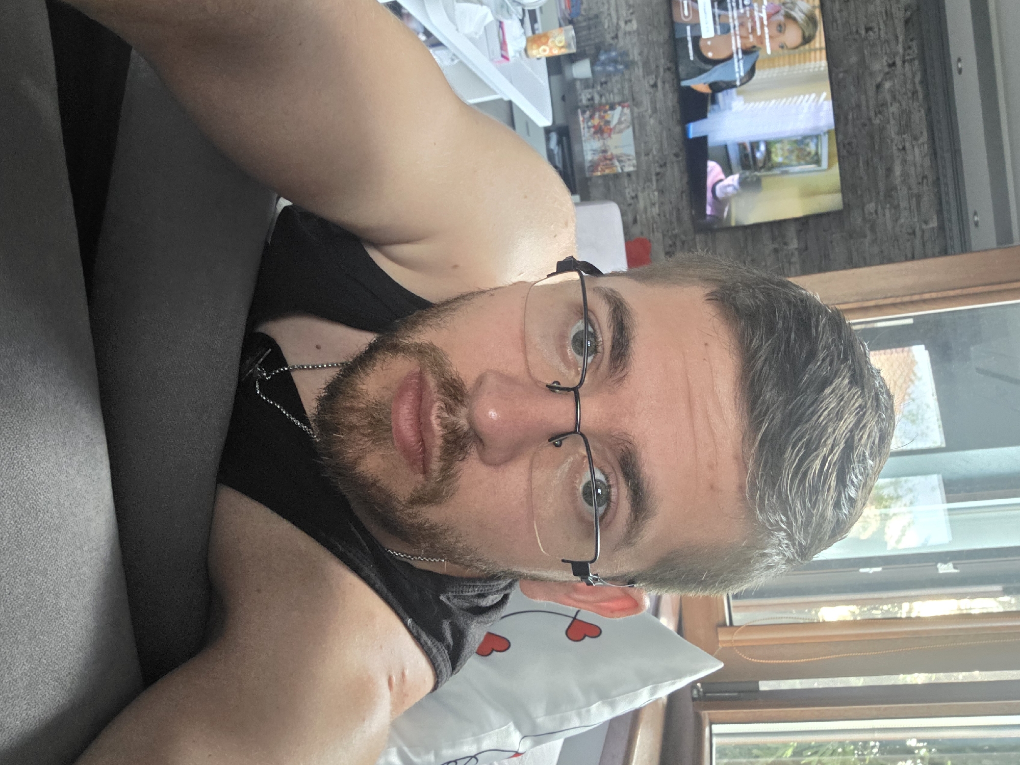 A man is reclining, facing the camera. He has a beard, wears glasses, and a black tank top with a silver chain. He appears to be on a gray couch, with a white pillow behind his head that has red hearts on it. Behind him are windows with blinds, offering a view of trees. In the background, further away, there's a wall with a television displaying a program. There is no visible evidence that places this image in Yagodovo, Bulgaria.
