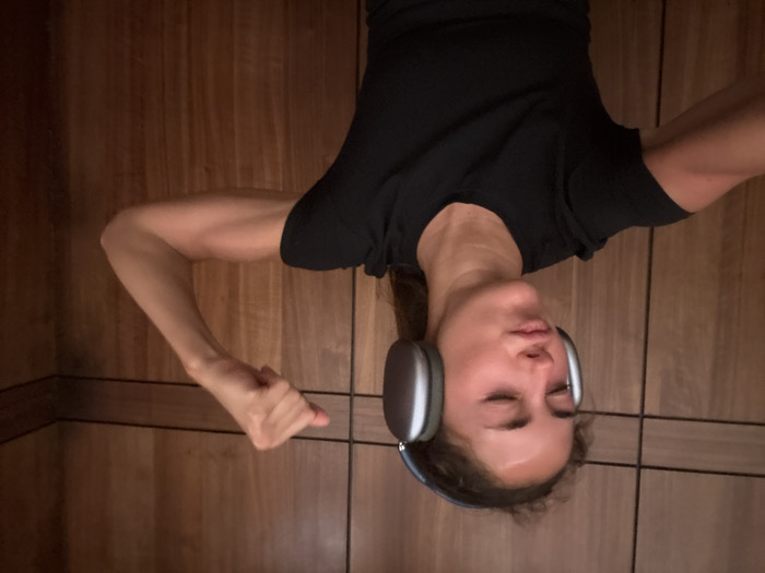 A woman hangs upside down, wearing headphones and a black t-shirt. Her eyes are closed. She is in a wood paneled sauna. Her arms are outstretched, one with a clenched fist. The background consists of vertical wood panels.