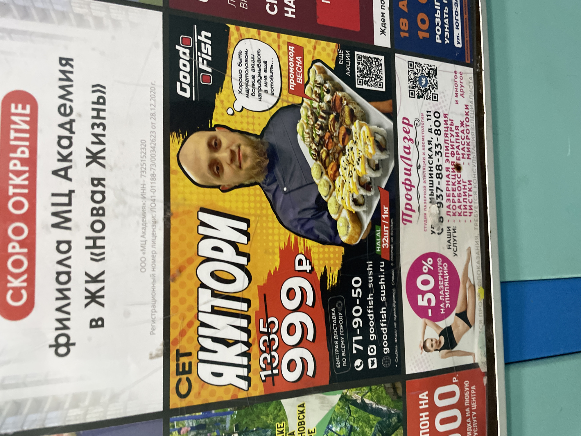 This media file displays a cluttered section of a wall covered with several overlapping advertisements in Russian, suggesting an indoor public or commercial space in Ulyanovsk, Russia.
The most prominent advertisement, positioned centrally, is for "Goodo Fish / ЯКИТОРИ" sushi delivery. It features a smiling bald man with a beard, presumably a chef, holding a large platter of sushi rolls. A humorous thought bubble above him reads, "It's good to be a marketer, they come up with all sorts of promotions, and I still have to cook...". The ad promotes a "SPRING" promo code, offering a sushi set for 999 ₽ (down from 1335 ₽), emphasizing "FAST DELIVERY ACROSS THE CITY," and listing a phone number (71-90-50), social media handles (@goodfish_sushi), and a website (goodfish_sushi.ru). It also notes the product is "HALAL" and includes "32 pieces / 1 kg," with a QR code for "MORE PROMOTIONS."
To the left, a vertical white advertisement announces the "COMING SOON" opening of a branch of "MC Academy" in the "New Life" Residential Complex, providing its TIN and license registration number from 2020.
On the right side, another advertisement is for "ПрофиЛазер" (ProfiLaser), a "STUDIO OF LASER EPILATION AND COSMETOLOGY" located at MyshinSKAYA, d. 111. It features a woman in black underwear demonstrating "-50% ON LASER EPILATION." The ad lists various services including figure correction, carboxytherapy, peeling, massage, microcurrents, and cleanses, along with a phone number (8-937-88-33-800) and a disclaimer about contraindications requiring specialist consultation. A QR code is also visible on this ad.
A partially visible ad at the bottom left offers a "VOUCHER FOR 1000 P." as a "DISCOUNT ON ANY SERVICE OF THE CENTER." At the top right, another incomplete ad mentions "18 A," "10," "DRAW," "FIND OUT," and "Yugo-Za street," suggesting a lottery or event.
The overall scene captures a vibrant marketing display, typical of a bustling urban environment, utilizing both visual appeal and informational text to attract customers to various local businesses. The lighting is bright, likely artificial.
