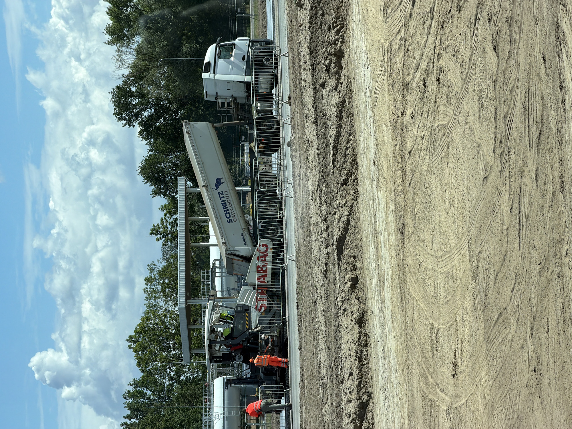 In Krzeptów, Poland, on a construction site, a road paving operation is underway. A paving machine, painted white with red lettering that reads "STRABAG", is actively laying down asphalt. A large white dump truck, identified as "SCHMITZ CARGOBULL", is positioned behind the paver, its bed tilted upwards, discharging material. Two construction workers are present: one in an orange high-visibility suit stands near the rear of the paver, and another in an orange vest and pants is on the left side of the paver, observing the process. A metal barricade separates the active work area from the surrounding ground. The environment is characterized by a partially constructed road surface, with areas of loose soil and tire tracks indicating recent activity. In the background, trees and a covered structure are visible under a bright blue sky with scattered white clouds.