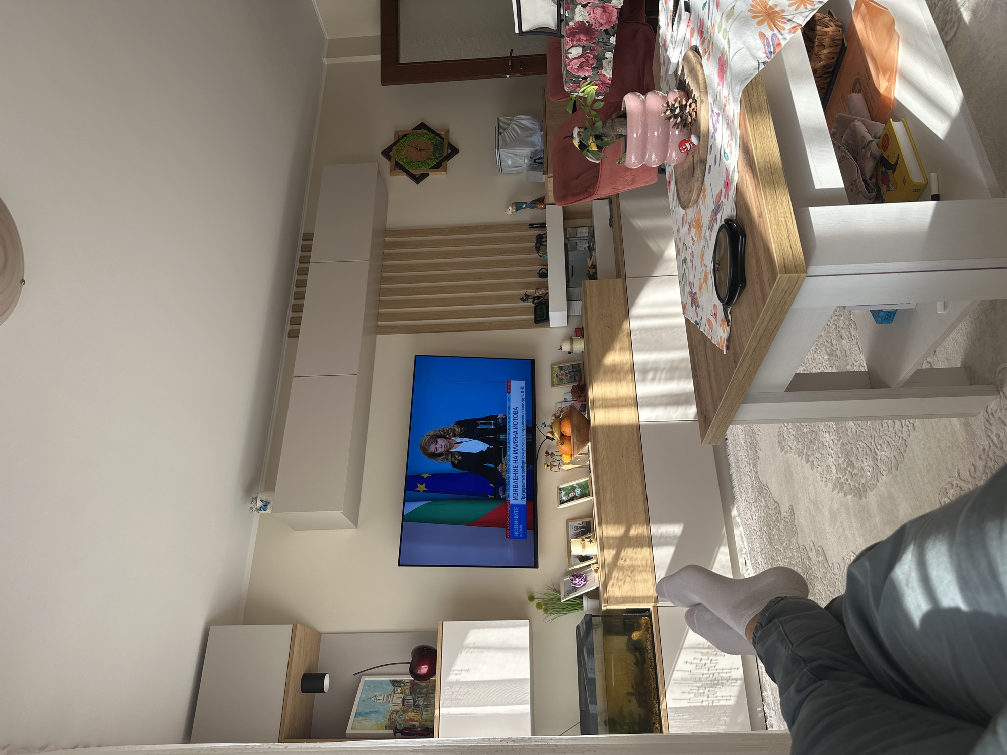 The image presents a residential living room interior. A television is mounted on the wall, displaying Bulgarian news. The decor features a combination of modern white storage units and natural wood accents. There's an aquarium positioned near the TV stand. A section of a person wearing gray pants and white socks is visible in the foreground. Additionally, part of a dining table with floral patterned linens and a red sofa are in view. The location is likely in Bulgaria given the language on the TV and the Bulgarian flag. However, the specific city is not identifiable from the details presented in the image.