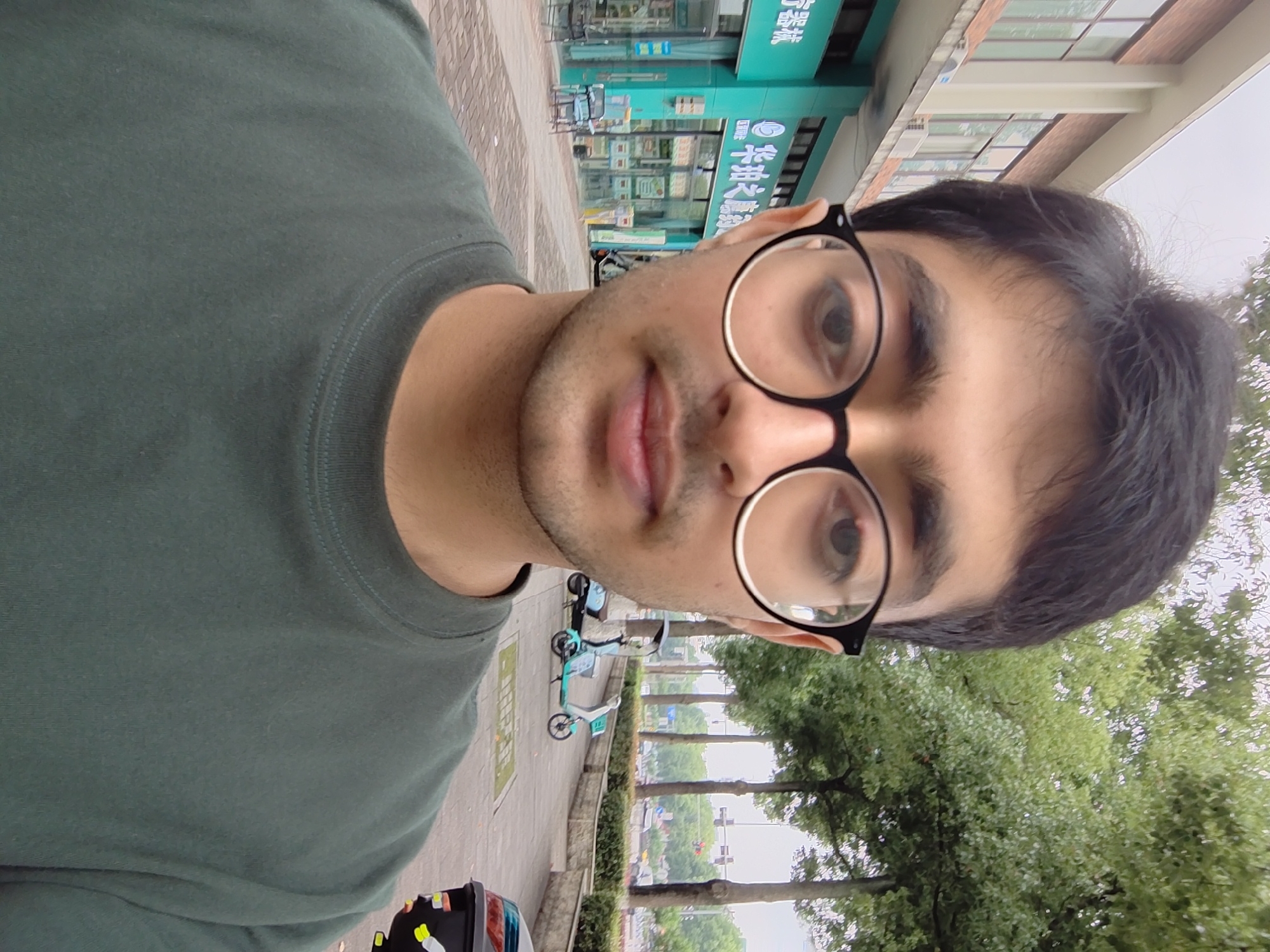 A young man with dark hair and glasses is taking a selfie outdoors. He is wearing a dark green crew-neck t-shirt. Behind him, a turquoise building with glass windows and signage in Chinese characters is visible. The building appears to be a shop or business. A sidewalk with a brick pattern runs in front of the building, and several parked electric scooters are present. Trees line the sidewalk, and in the background, a street with traffic and traffic lights can be seen. The sky is overcast. The location is identified as the Chongqing High-tech Zone in China.