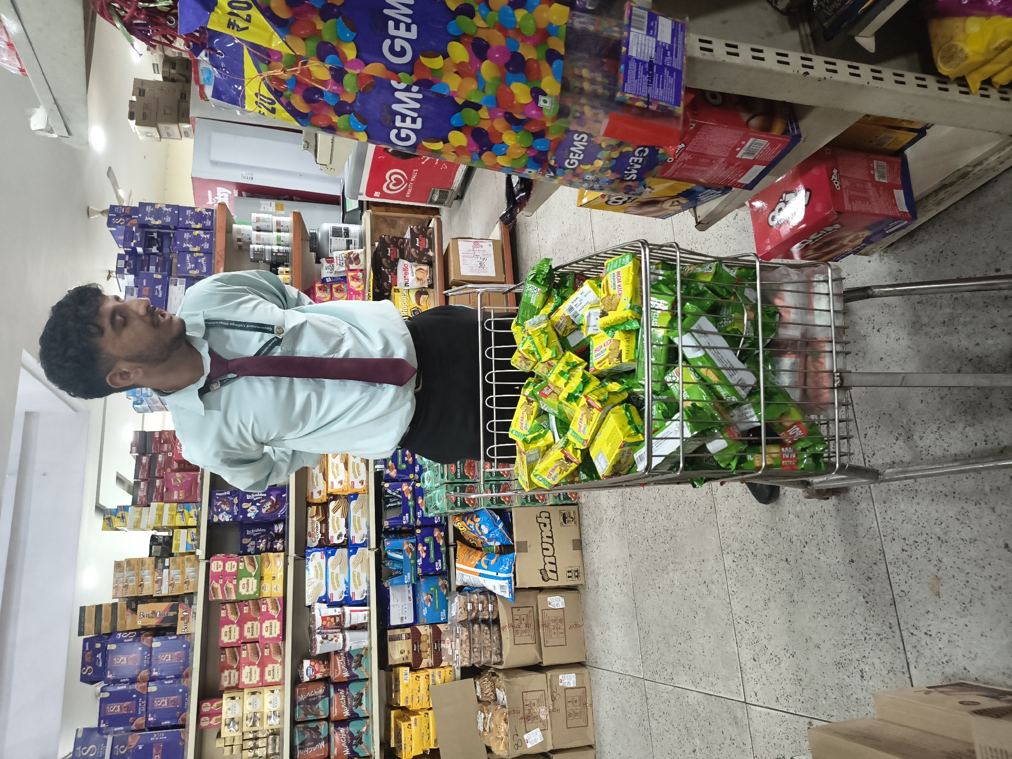 In a retail store in Sidhpur, India, a young male employee wearing a light blue shirt and a maroon tie stands near a shopping cart overflowing with green-packaged snacks. The cart's contents appear to be individual snack portions, possibly biscuits or wafers, with some portions of larger snack bags also visible at the bottom. Behind the employee, shelves are stocked with various consumer goods, including boxes of chocolates, packaged snacks, and other food items. A large display of colorful candies in transparent packaging is situated to the right of the employee. The store has a tiled floor and fluorescent lighting, typical of a supermarket or convenience store environment. The employee is looking towards the shopping cart and appears to be overseeing the stocked items.