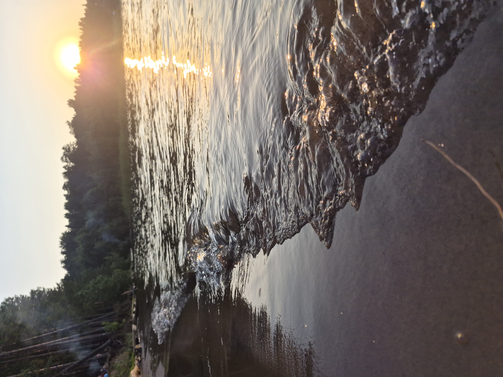 A low-angle, close-up shot captures a gentle wave lapping at the dark, sandy shore of the Kama River at sunset in Chaikovsky, Russia. The setting sun, a bright orb low on the horizon, casts a golden sheen across the rippling water, creating a shimmering path of light that extends towards the viewer. Silhouetted trees form a dense forest along the far bank, with a hint of smoke rising from what appears to be a campfire among them, suggesting the presence of people. The water is clear, with small whitecaps and bubbles visible as the wave breaks. The sand is dark and wet, reflecting the soft light of the sunset.