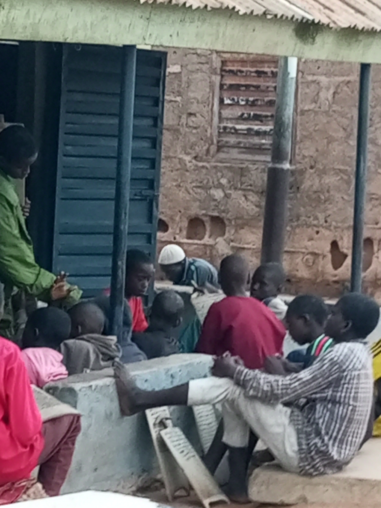 In Garko, Nigeria, a group of children and an adult are gathered outside a building with a corrugated metal roof and plastered walls. The adult, wearing a green uniform, stands near a dark blue shuttered doorway, appearing to be distributing something held in a green cloth. The children are seated on a concrete structure. One child in the foreground wears a pink top and maroon pants, while another to their right has on a red shirt. Further to the right, a child in a checkered shirt and white pants is seated. Several other children are partially visible in the background. One of the children in the background wears a white skullcap. The environment appears to be an outdoor setting, possibly a community center or school.