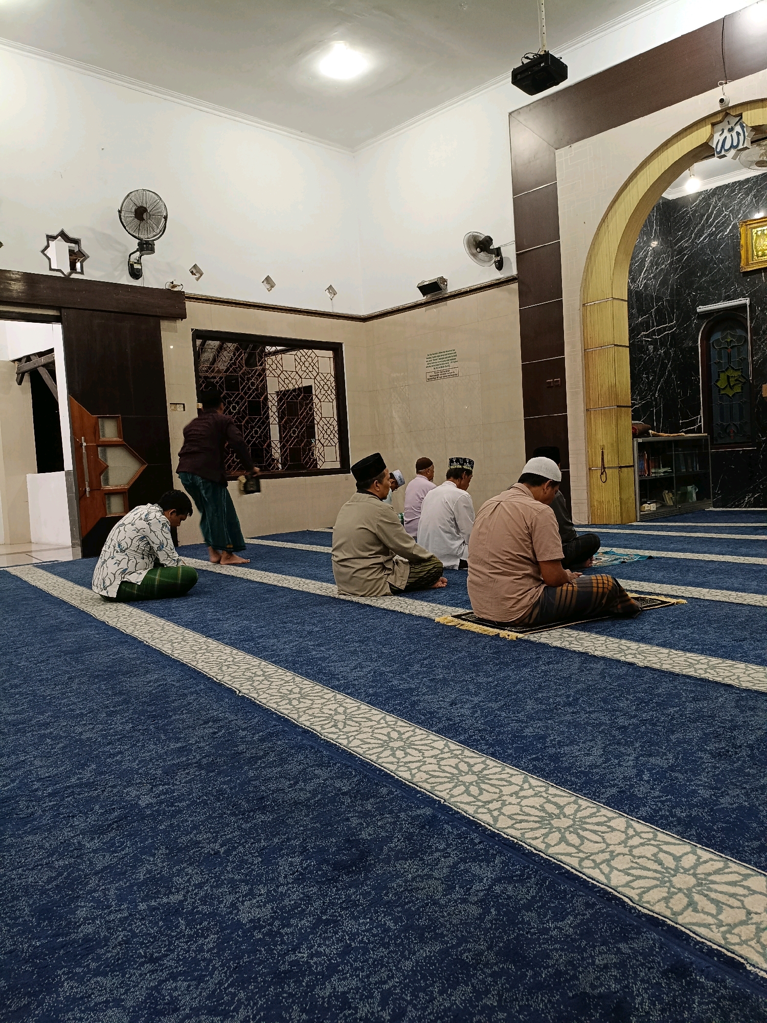 In the city of Maguwoharjo, Indonesia, a mosque interior is depicted, featuring a group of men engaged in prayer. The flooring is a deep blue carpet with decorative white and grey patterned strips. Several men are seated on prayer mats, facing forward in unison. One individual, dressed in a patterned shirt and green sarong, is seated on the floor with his back to the camera. Another man in a light brown shirt and checkered sarong is kneeling on a prayer mat. Further back, two men in white shirts and head coverings are also seated. A fifth man, in a dark maroon shirt and green pants, stands near a decorative window, holding a book. The walls are a light cream color, with some sections featuring dark brown paneling and an arched doorway leading to a marble-patterned area. A wall-mounted fan is visible on the left side of the room. Overhead, ceiling lights illuminate the space. The overall atmosphere suggests a moment of quiet devotion within a religious setting.