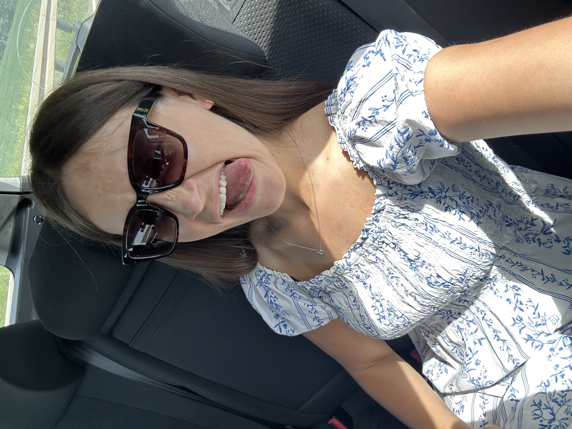 A young woman is in the back seat of a car, wearing sunglasses and a white, floral print dress. She is sticking her tongue out and making a face. The background suggests the interior of a vehicle, with parts of seats visible. Based on the geographical implications from the prompt, the location is inferred to be the city of Lich, Germany, though there's no confirmation of this within the image itself. No specific actions, interactions, or particular environmental cues apart from the interior of a car are discernible.