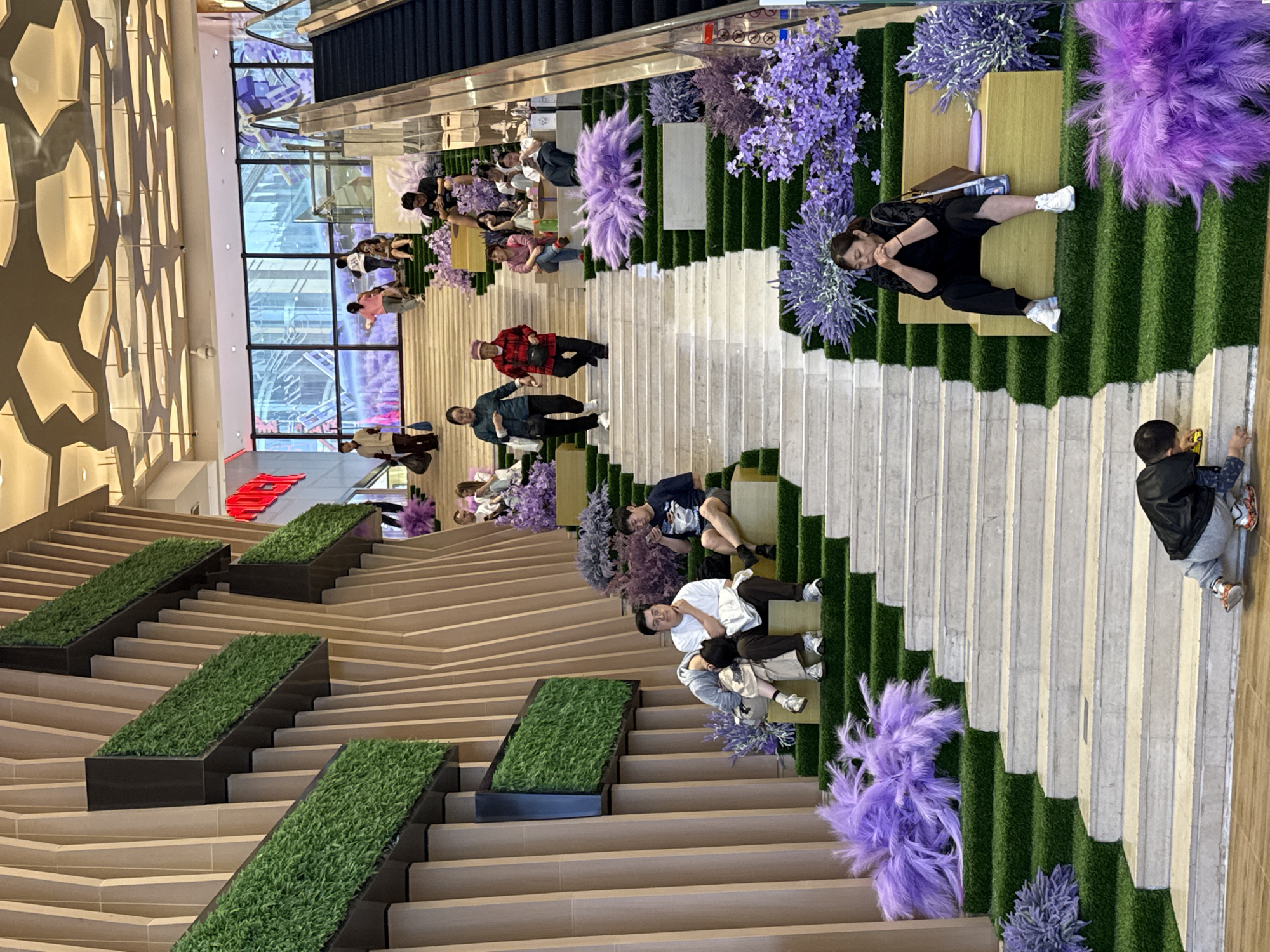 The image shows a multi-level indoor space, likely a shopping mall, in Yuzhong District, China. The space is designed with a modern, stepped platform style. People are seen sitting and resting on various levels of the platform. There are decorative elements, including greenery in rectangular planters and purple floral arrangements, lining the platforms. An escalator is visible in the upper part of the image, and a glass wall allows natural light into the building.