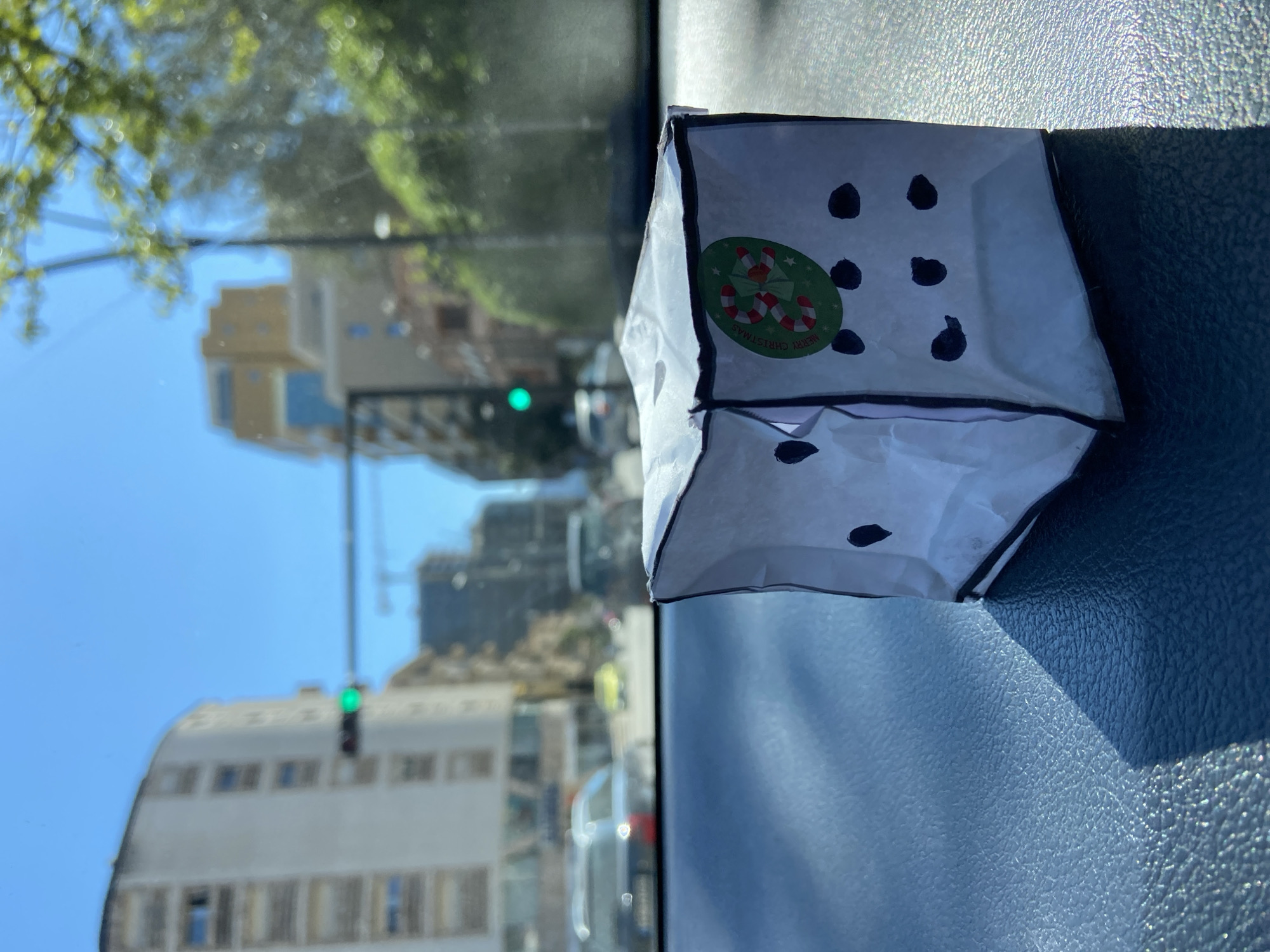 The image shows a small, white paper bag designed to resemble a die sitting on a dark gray textured surface, likely a car dashboard, with a green "Merry Christmas" sticker affixed to one side. The die design is depicted with black dots. The background, visible through the car's windshield, features a blurred urban cityscape with buildings, a green traffic light, and foliage. The exact location in Budva, Montenegro is not discernible from the image alone, but the presence of urban buildings and a green traffic light suggest a street scene within the city. No people are directly visible in the image.