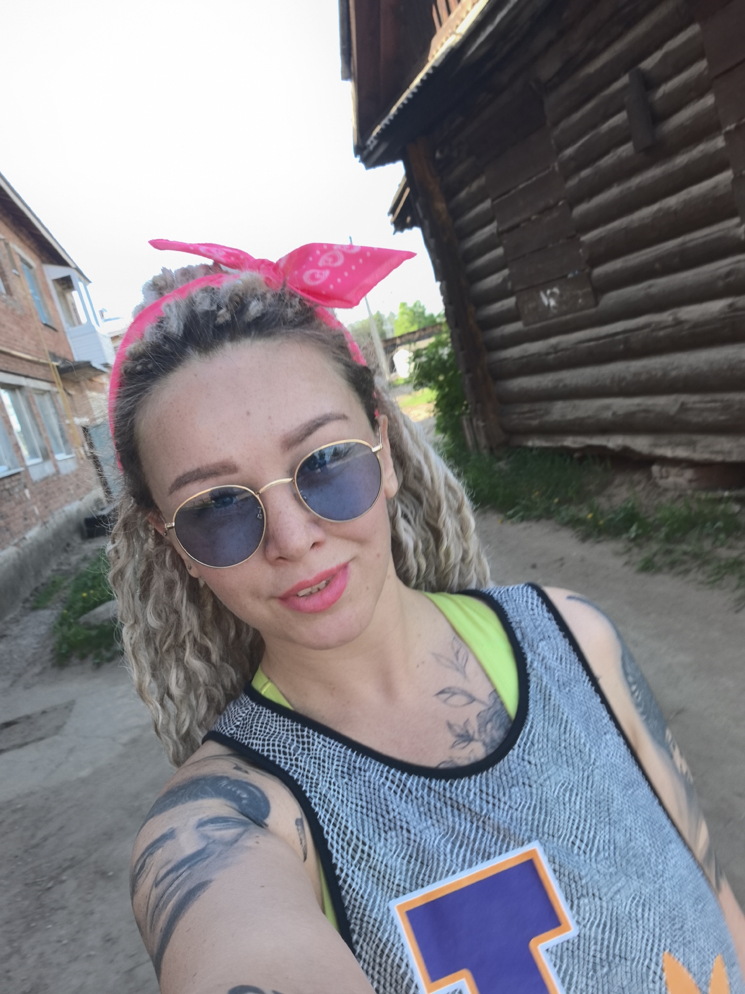 A medium shot of a light-skinned woman with blonde curly hair wearing round sunglasses and a pink bandana. She is wearing a gray tank top over a neon yellow shirt. Visible tattoos adorn her arms and chest. She appears to be on a dirt road in Noviy, Russia. To the left is a two-story brick building with windows. To the right is a log cabin with visible moss. The lighting indicates it's daytime.