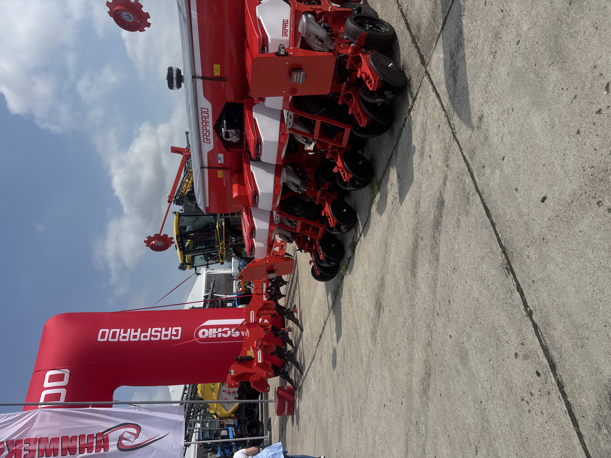 This media file captures an outdoor exhibition of agricultural machinery in Stara Zagora, Bulgaria, under a bright, partly cloudy sky. The primary subject is a large, multi-row, red and white agricultural planter or seeder, branded "GASPARDO" and "MASCHIO," which extends horizontally across the central and right portions of the frame. This piece of equipment features numerous planting units, each with disc openers and various mechanical components designed for sowing.
To the left of the main machinery, a large, red inflatable archway is visible, also prominently featuring the "MASCHIO GASPARDO" brand name (which appears upside down relative to the current image orientation). A partially visible yellow tractor is connected to or positioned near the planter, suggesting its function as a towed implement. Further in the background, other exhibition tents and machinery can be glimpsed.
The ground is concrete, showing distinct seams and some shadows, indicating it is daytime. A person wearing a light blue shirt is partially visible at the bottom-left edge of the frame, hinting at visitor presence. A white banner with partial, reversed text "HHNWEK" is also visible at the bottom-left. The scene suggests an active agricultural trade show or fair, showcasing modern farming equipment.