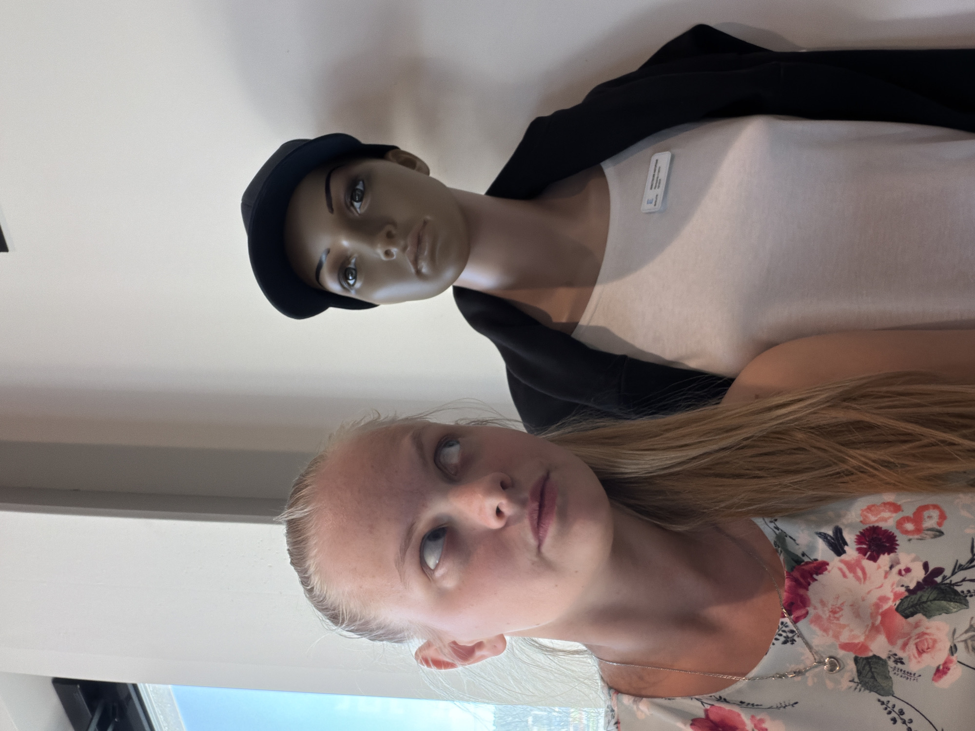 The image shows a fair-skinned young woman with blonde hair posing next to a mannequin wearing a white shirt, black jacket, and black hat. The woman is wearing a floral-patterned dress and a silver necklace. The scene appears to be indoors, possibly in a retail or display setting. The location in Izhevsk, Russia, is not definitively indicated in the image.