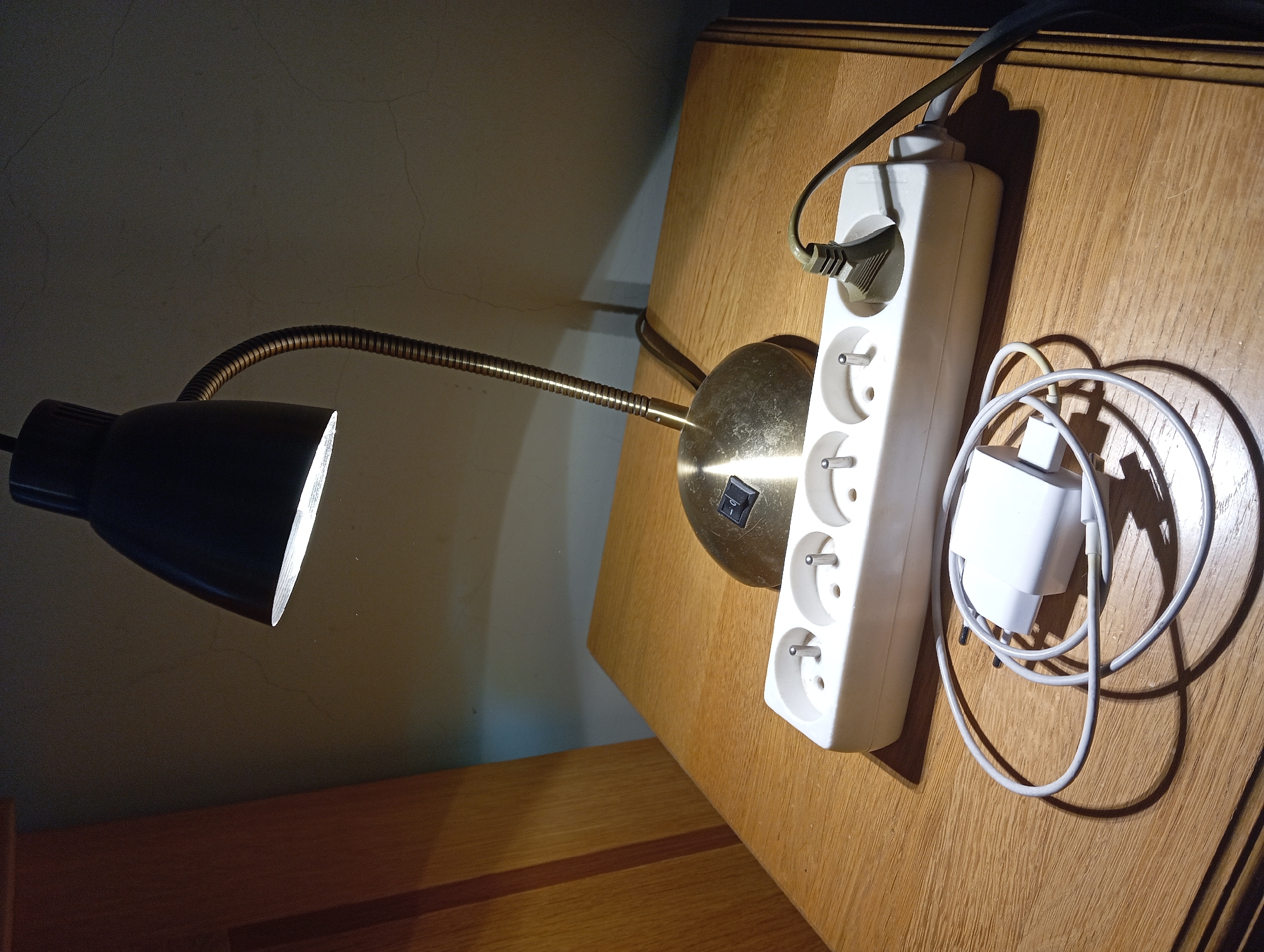 The image depicts an indoor scene in Nitra, Slovakia. A black desk lamp with a brass-toned gooseneck stem and base is actively lit, projecting illumination downwards onto a light brown wooden surface. The lamp's dark green Type F power plug is inserted into the rightmost socket of a white four-outlet Type F power strip. Positioned on the wooden surface, immediately in front of the power strip, is a white mobile device charger consisting of a USB power adapter and a coiled white cable. The wooden surface displays visible grain patterns, consistent with a desk or nightstand. In the background, a light-colored wall with multiple fine cracks is visible, partially illuminated by the lamp. To the left, a segment of vertical wooden furniture is present, matching the tone of the horizontal surface. The scene's primary light source is the desk lamp, creating distinct shadows from the objects.