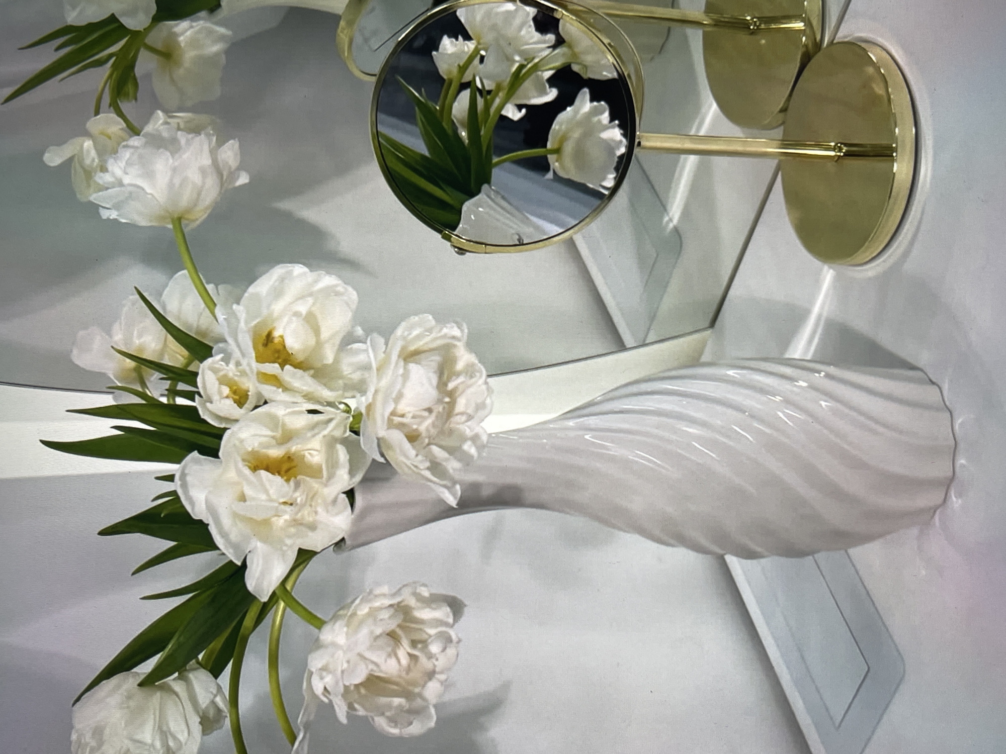 This media file displays a close-up, eye-level shot of a white vase holding a bouquet of white tulips. The vase is made of glass with a swirling, ribbed texture and is positioned on a white surface. The tulips are fully bloomed, with layered petals and prominent yellow centers. Green leaves and stems are also visible. In the background, a round mirror reflects a portion of the tulips and stems, adding depth to the composition. The mirror is framed in gold and appears to be attached to a wall-mounted fixture, also in gold. The lighting is soft and diffused, suggesting an indoor setting with ample natural or artificial light. The overall mood is serene and elegant, emphasizing the natural beauty of the flowers and the design of the vase and mirror. There is no visible text in the image.