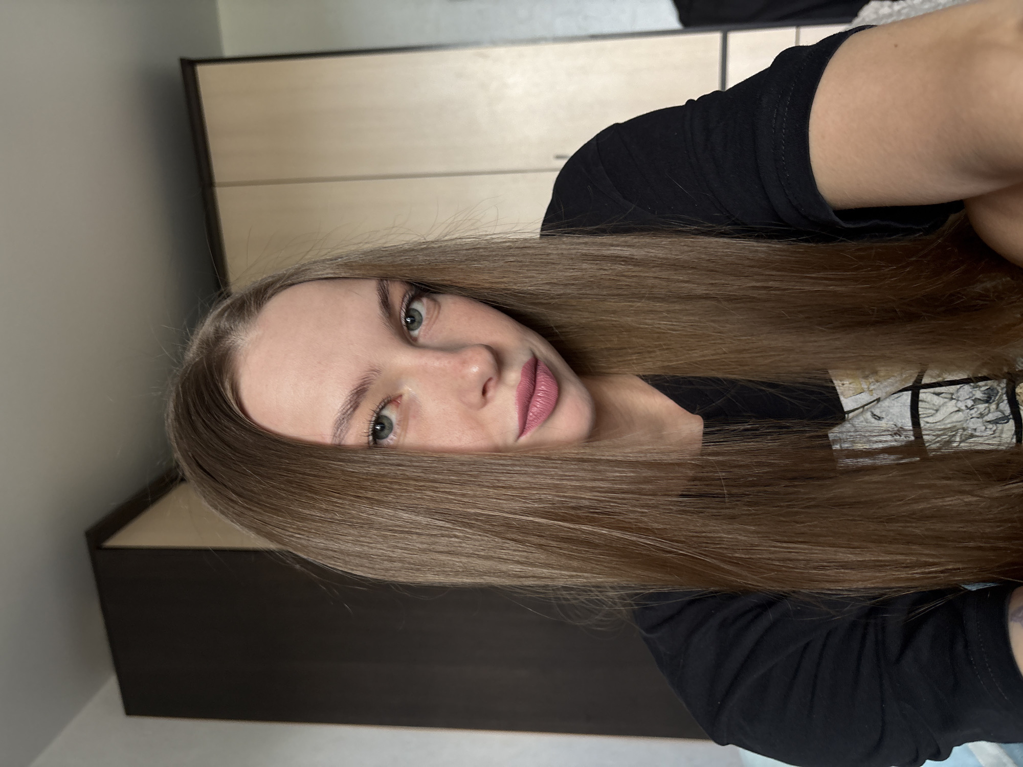 The image shows a young woman with long, brown hair and fair skin, wearing a black t-shirt. She is looking directly at the camera with green eyes and pink lipstick. The background appears to be an indoor setting, possibly a bedroom, with a closet or wardrobe visible. There is no discernible connection to the city of Perm, Russia.