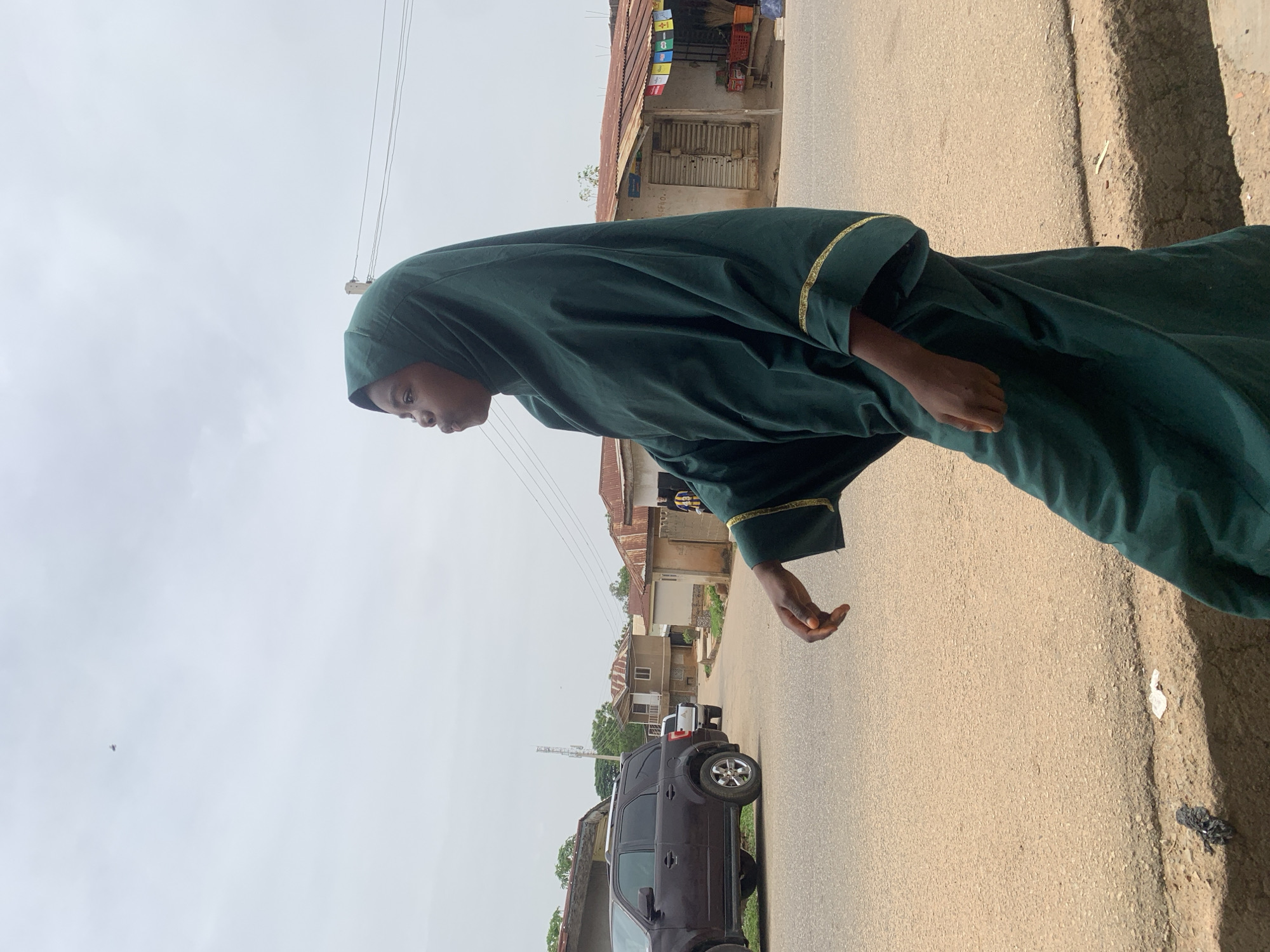 A person wearing a dark green hijab-like garment with gold trim stands on a paved road. The person is facing left and is slightly angled towards the camera.
The background includes buildings and cars. A dark SUV is parked on the side of the road. The buildings appear to be a mix of residential and commercial structures. A storefront with flags hanging outside is visible in the background. The sky is overcast.
The location appears to be in Jos, Nigeria, given the architecture and environment.