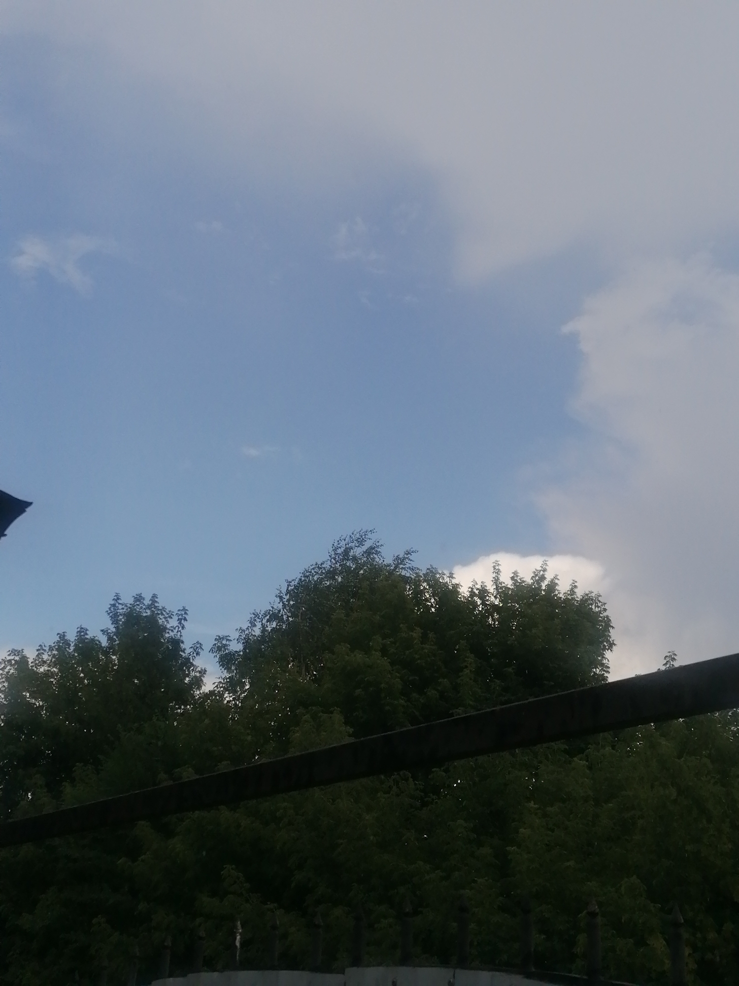 The image shows a slightly overcast sky with patches of blue and white clouds. The top of a dark structure, possibly a roof or a large object, is visible in the upper left corner. A thick, dark horizontal beam or pipe runs across the middle of the frame. Below this beam is a dense canopy of dark green trees. In the foreground, a dark fence with pointed posts is visible. The scene appears to be an outdoor view, possibly from an elevated position, looking over trees towards the sky. The location is the city of Serebriansk, Kazakhstan.