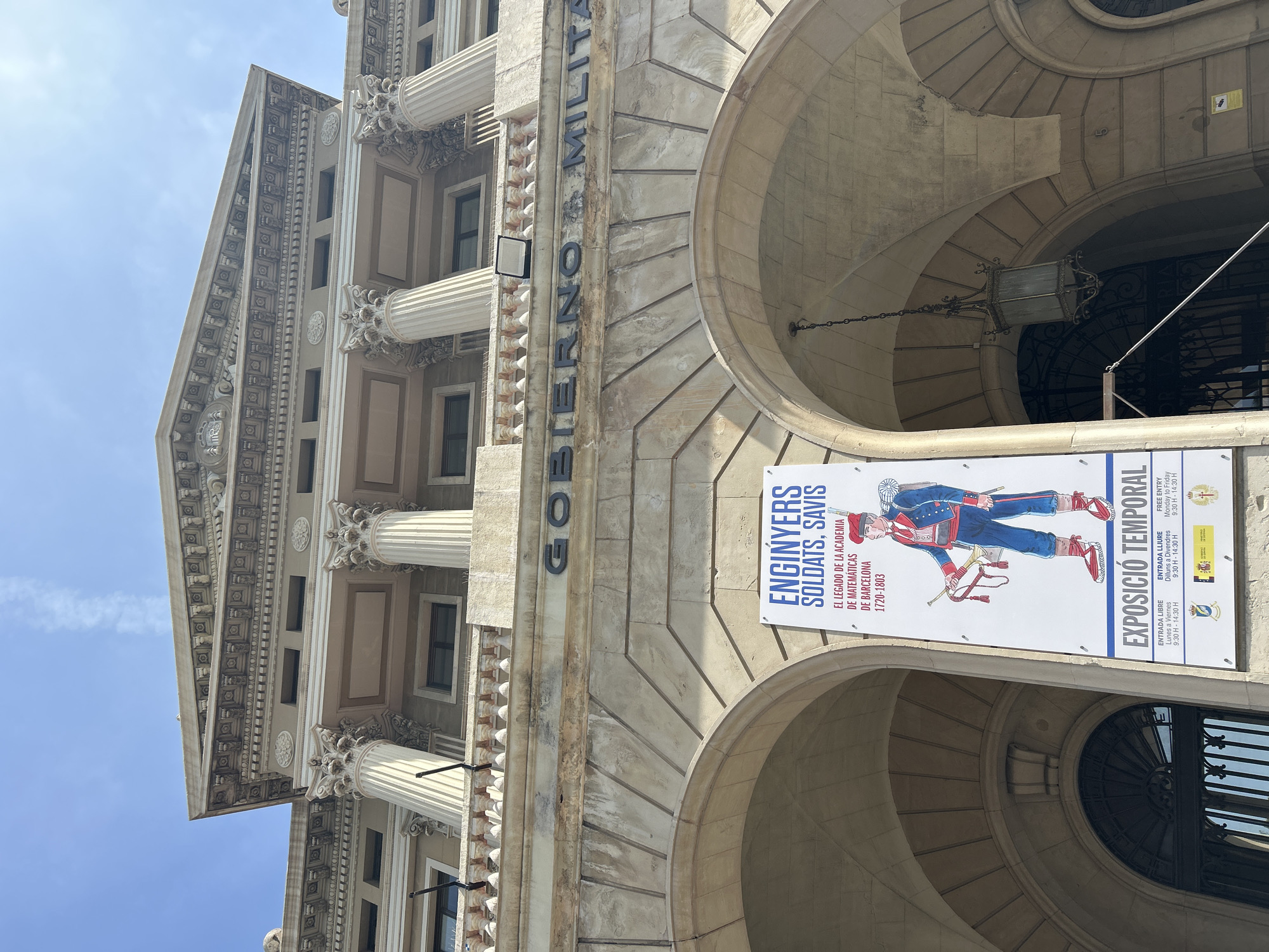 The image shows the facade of a building in Barcelona, Spain, with a poster announcing a temporary exhibition titled "Enginyers Soldats Savis" (Engineers, Soldiers, Wise Men) which covers the legacy of the Academy of Mathematics of Barcelona from 1720 to 1803. The poster also indicates the exhibition is free and lists the opening hours from 9:30 AM to 2:30 PM, Monday to Friday. The building itself is of classical design, featuring columns, arches, and decorative elements. The words "GOBIERNO MILITA" are written vertically on the side of the building.