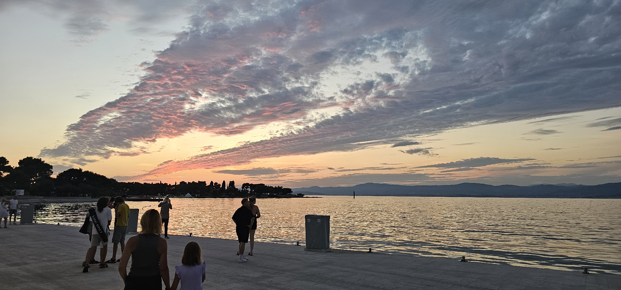 The image depicts a sunset over the sea in Grad Supetar, Croatia. A group of people are standing on a paved promenade. The sky is filled with dramatic clouds, tinged with pink and orange from the setting sun, contrasting with areas of grey and blue. The sea reflects the colors of the sky, with gentle ripples on its surface. In the distance, a tree line borders the coast, and mountains are visible on the horizon. The overall scene is serene and picturesque, capturing the beauty of a coastal sunset.