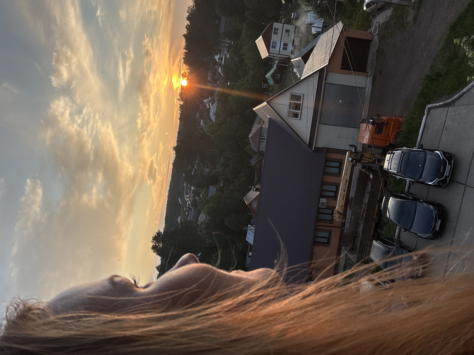 The image captures a sunset over a semi-rural area in Chaikovsky, Russia. In the foreground, the side profile of a person with blonde hair is visible, looking out towards the horizon. Their face is partially obscured by their hair, but their eye and the bridge of their nose are discernible.
The midground of the image features several houses and buildings, characteristic of a suburban or village setting. A prominent dark-tiled roof of a larger building dominates the left side of this area. To the right, a two-story house with a light-colored facade and dark roof is visible. A bright orange truck is parked near a building with a large garage door. A crane is also visible, suggesting construction or maintenance work.
The background is filled with a dense forest that extends to the horizon. The sky is a canvas of warm colors, with the sun low on the horizon, casting a radiant glow and visible sun rays. Clouds are scattered across the sky, tinged with orange and yellow hues from the sunset. Two cars are parked in a paved area in the foreground, to the right of the frame. One is a dark sedan, and the other is a darker SUV.