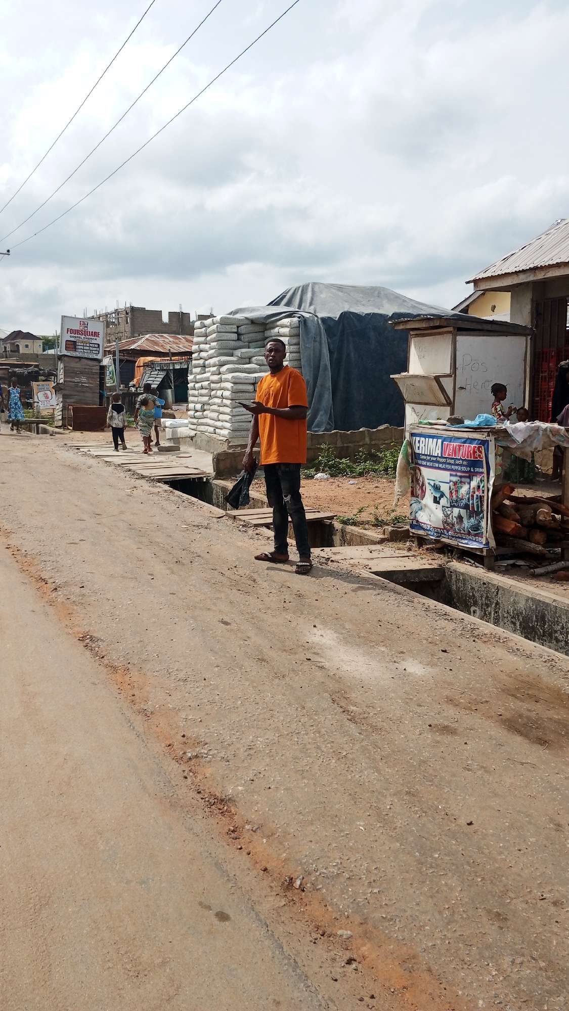 The image captures a street scene in what appears to be Zango-ndaji, Nigeria. The street is unpaved and sandy, with open drainage ditches running along the sides.
A man wearing an orange t-shirt and dark jeans stands in the middle ground, holding a mobile phone in his right hand and a black plastic bag in his left.
Several children are walking further down the street.
A stack of bags is visible behind the man, covered by a tarpaulin. Adjacent to this stack is a small kiosk with a hand-painted sign reading "Pos Here". Another sign, titled "KERIMA VENTURES", advertises pepper soup, including goat meat, cow meat, fish pepper soup, and drinks.
Signs for "FOURSQUARE" can also be seen.
The sky is overcast.
