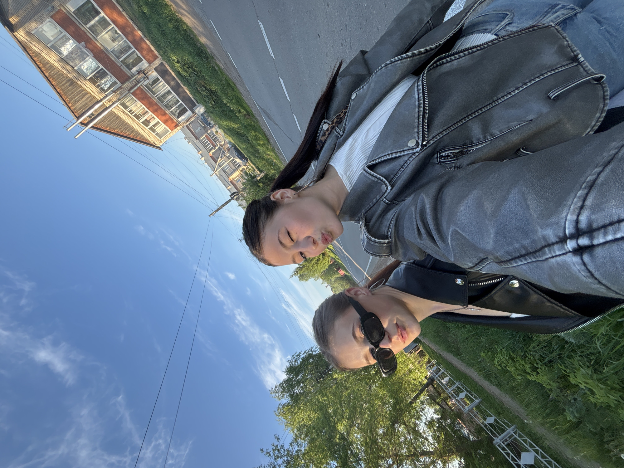 Two women are posing for a selfie on a road in Фоки, Russia. One woman wears a grey leather jacket and jeans. The other woman wears a black leather jacket and sunglasses. In the background, there is a blue sky with some clouds, trees, grass, a road with a white line, and a building. There are also power lines visible.
