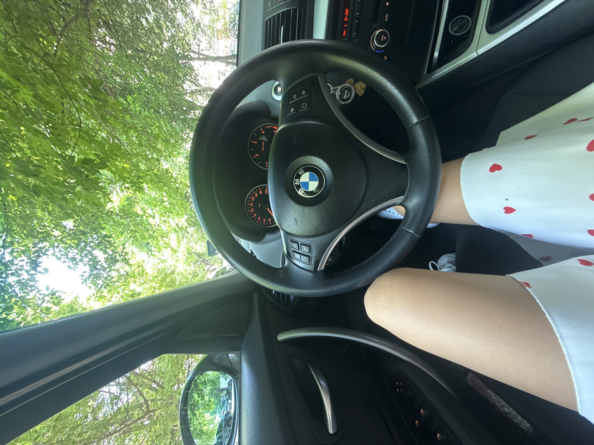 The image presents a first-person interior view from the driver's seat of a parked BMW vehicle in Plovdiv, Bulgaria. The driver's bare thighs are visible, along with a white garment patterned with red hearts covering the lower lap. White sneakers are partially visible on the floor. The steering wheel, bearing the BMW logo, is equipped with multifunction controls, and a keyring with a charm hangs from the steering column. The instrument cluster shows the speedometer and tachometer at zero, indicating the vehicle is stationary. The center console displays a radio tuned to "FM 6 N-JOY" and climate controls. The interior trim is dark, with metallic accents on door handles. Through the windshield and reflected in the side mirror, dense green tree canopies are visible, suggesting a tree-lined street or park area under bright daylight conditions. Parked cars are also reflected in the side mirror.