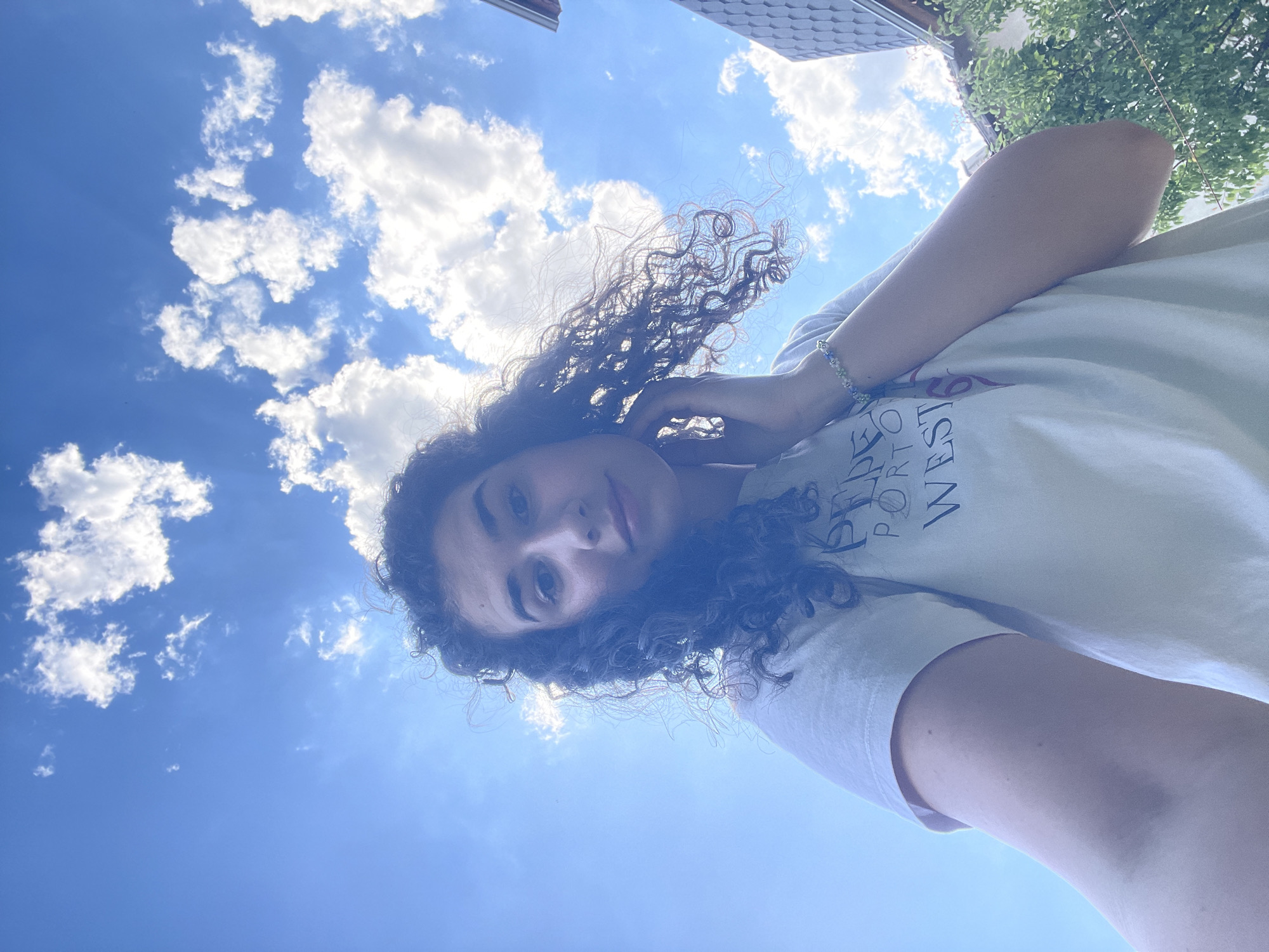 A young woman with curly dark hair poses for a selfie against a bright blue sky with scattered white clouds. The photograph is taken from a low angle, looking up at the subject. The woman wears a light-colored t-shirt with lettering visible on the upper portion of the garment. Her left arm is raised, with her hand near her face. A bracelet is visible on her left wrist. The background includes portions of buildings and trees. The buildings and trees are not detailed enough to pinpoint the precise location of Sofia, Bulgaria.