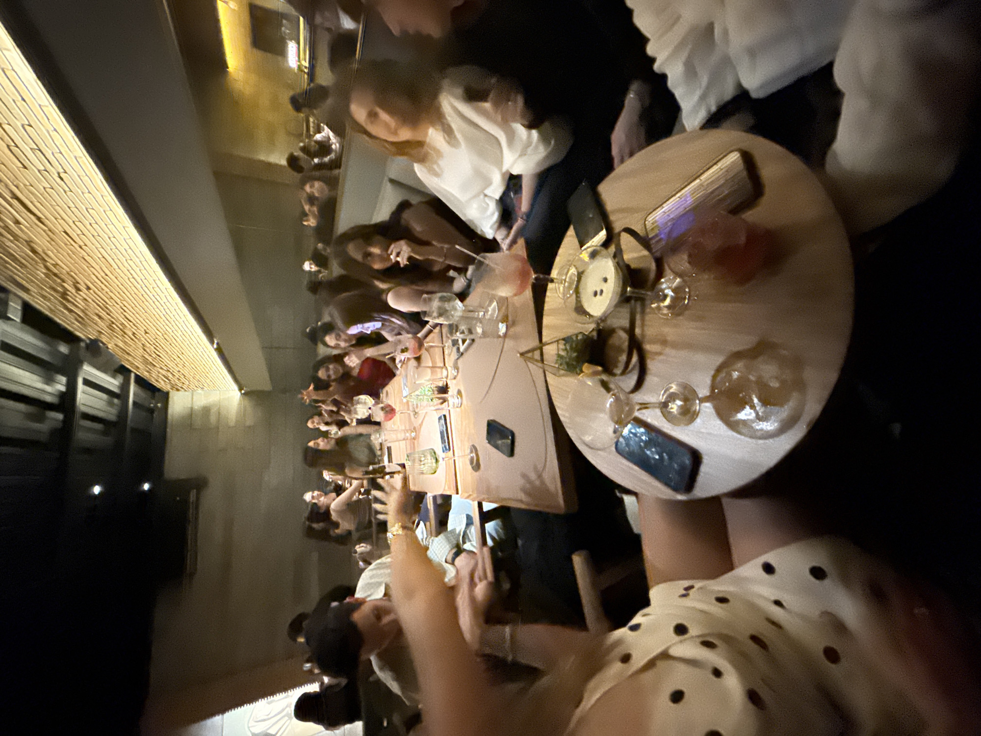 A group of people are seated at a table in what appears to be a restaurant or bar. The location is likely in San Andrés Cholula, Mexico. They are eating and drinking, with drinks and appetizers on the table, illuminated by overhead lighting. Some people are taking pictures with their phones. The group is having a social gathering or celebration.