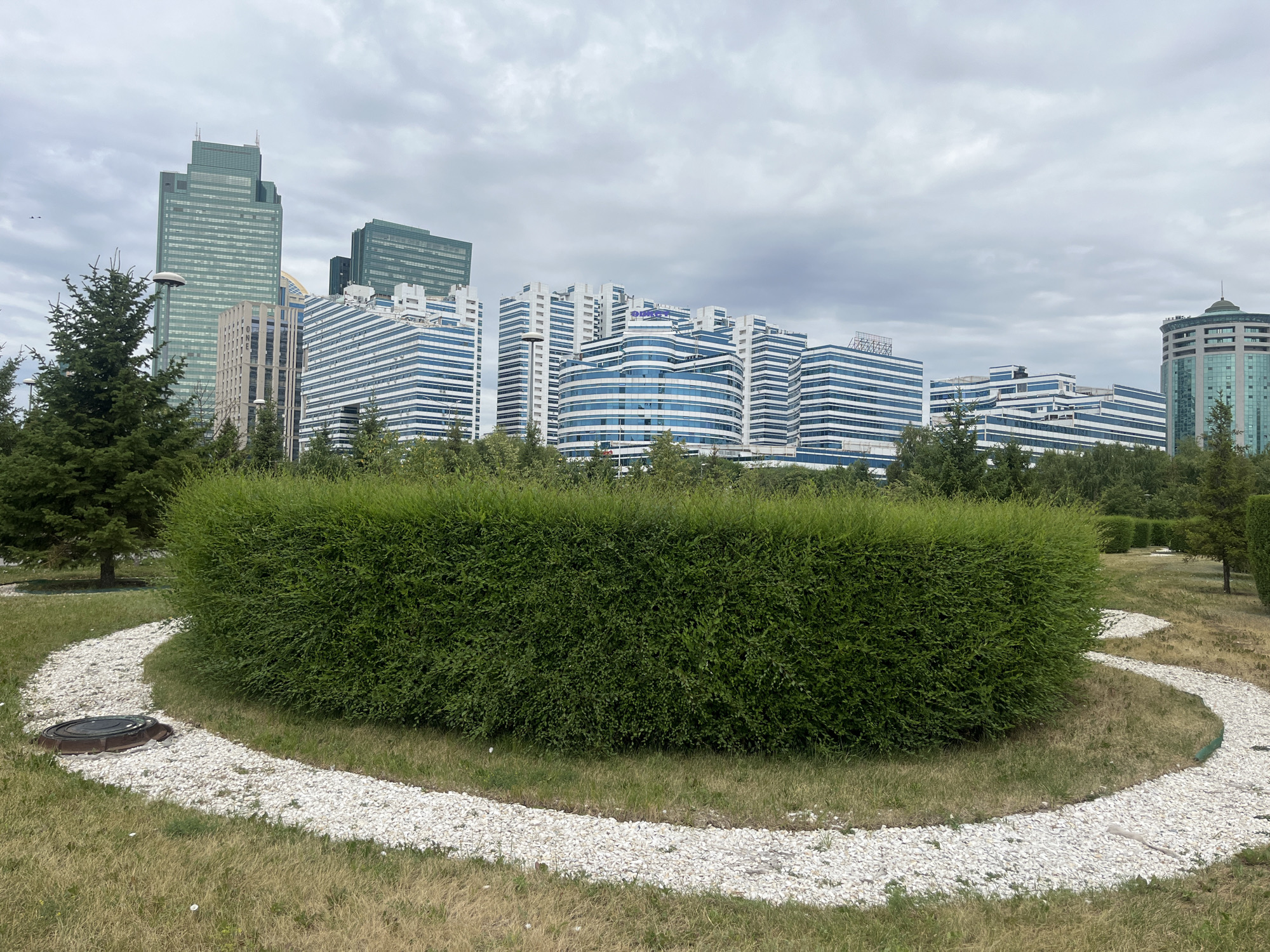 The image shows a city park in Astana, Kazakhstan, with modern skyscrapers in the background. In the foreground, a large, meticulously trimmed green hedge forms a central feature, surrounded by a winding path of white pebbles. A manhole cover is visible on the pebble path. The ground is covered with grass, some of which appears dry. Scattered trees, including coniferous species, are present in the park. The sky is overcast with clouds. The buildings in the background exhibit contemporary architectural styles, with many featuring glass facades and geometric designs. One prominent building appears to have a distinctive blue logo. The overall scene depicts a well-maintained urban green space contrasting with the imposing cityscape.