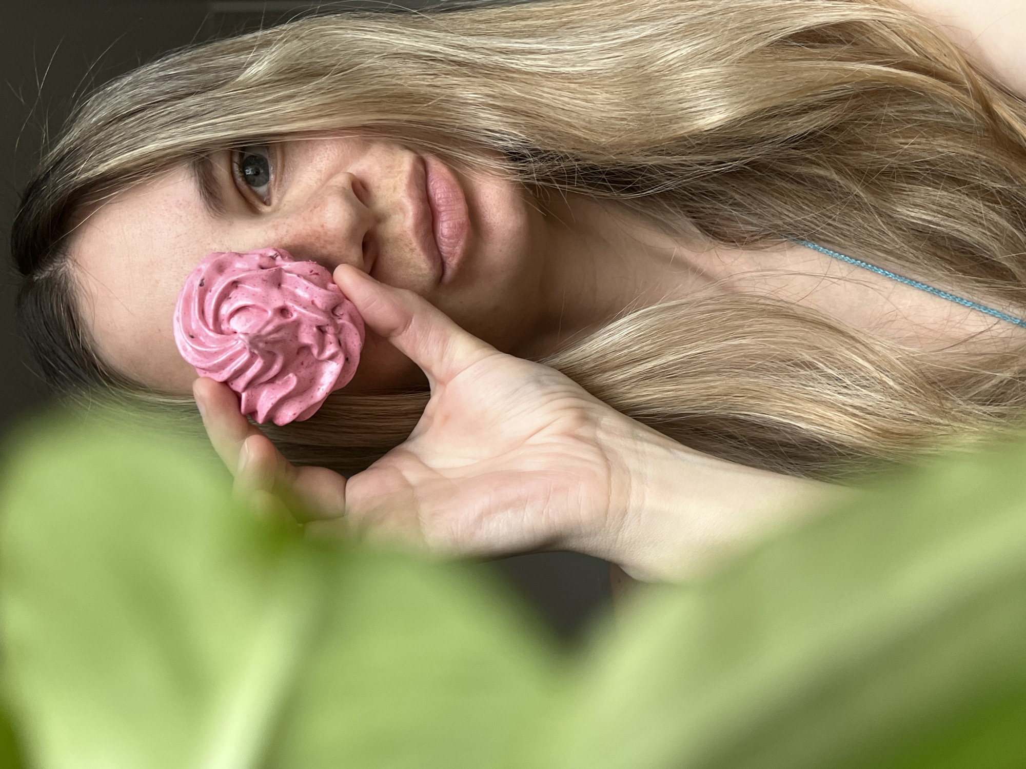 A woman is holding a small, pink, flower-shaped confection near her face. The confection appears to be a meringue. The woman has light blond hair and light skin. She is partially obscured by green foliage in the foreground. A thin blue strap is visible at her shoulder. The image appears to be taken at an unusual angle, rotated to the left. The location and time are not determinable from the given image.