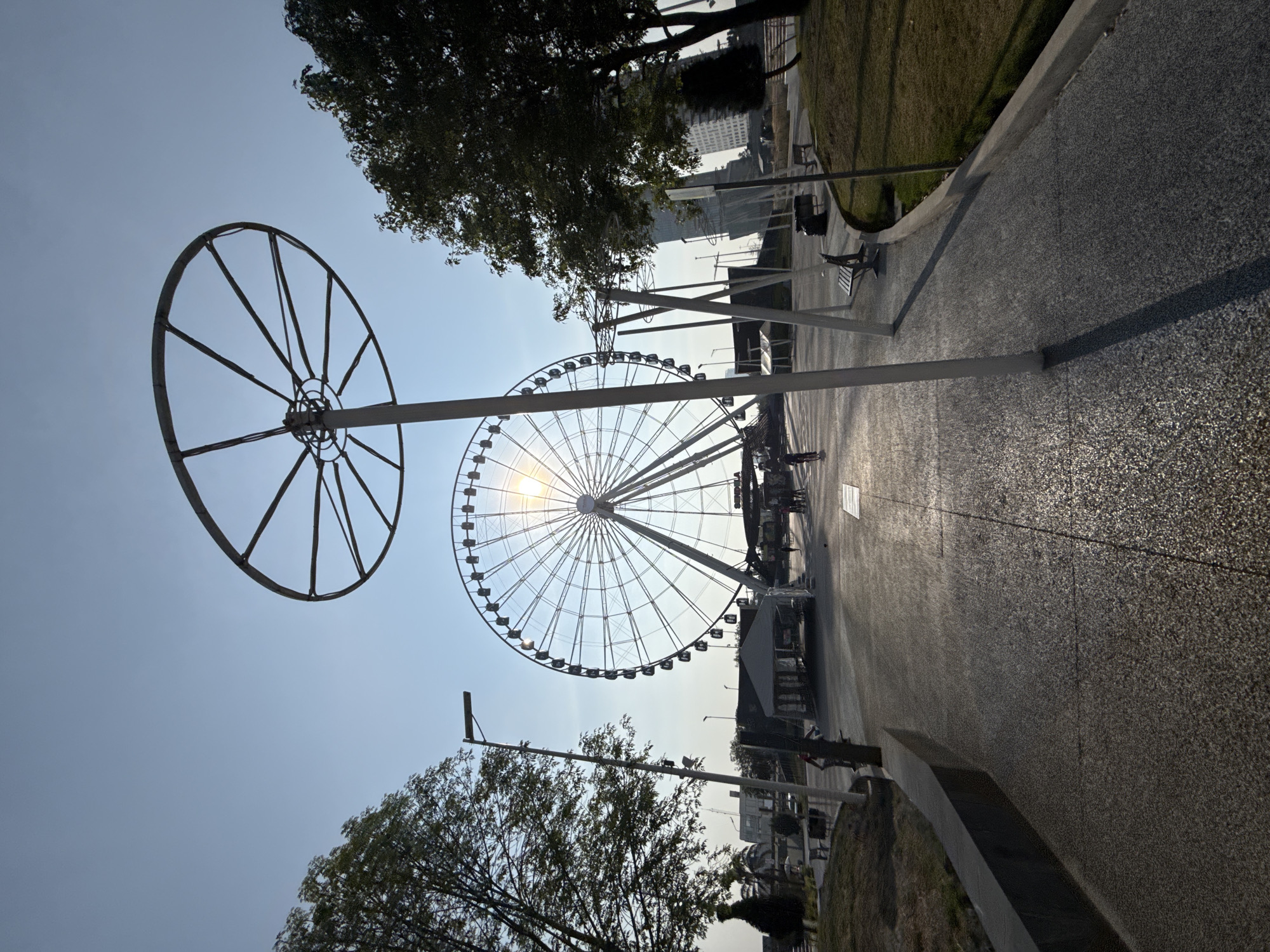 This is an outdoor shot of a park area in Puebla, Mexico, featuring the Estrella de Puebla Ferris wheel. The image is taken from a low angle, looking upwards at the ferris wheel, which dominates the scene. The sun is behind the Ferris wheel, creating a bright glare. To the left is a smaller, skeletal wheel-like structure mounted on a pole. Trees frame the bottom and top of the image. A paved pathway with benches and other park infrastructure extends into the distance. The pathway appears to be busy with people. The weather is sunny and the sky is clear. Buildings are visible in the background.
