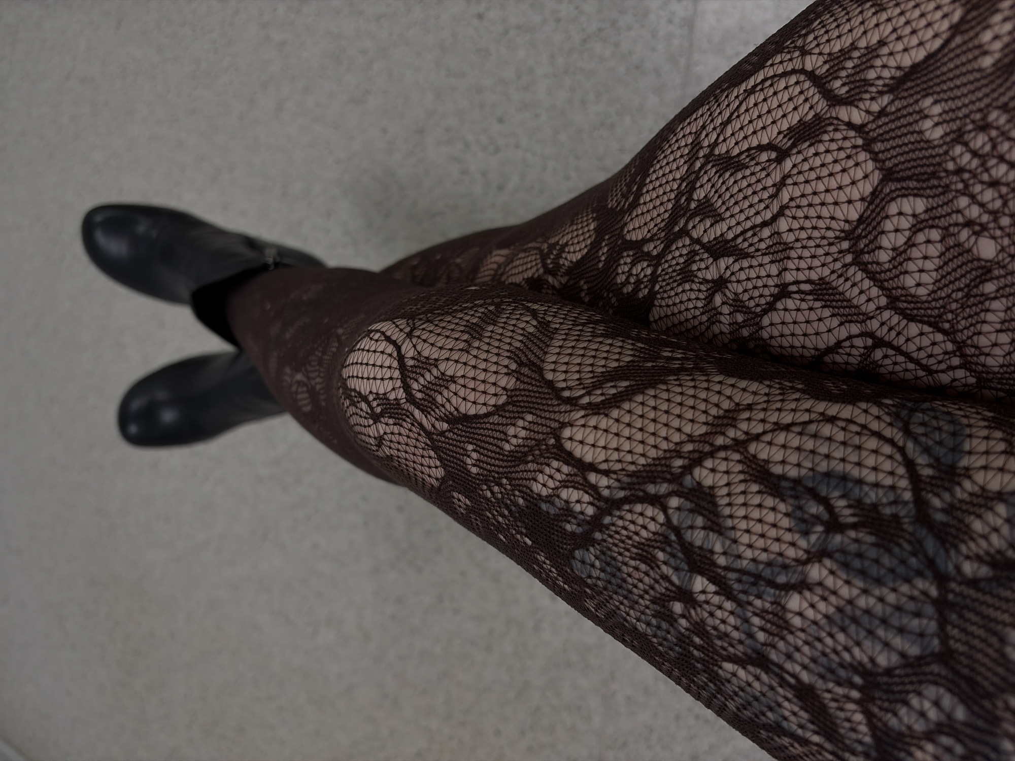 A close-up, downward-angled shot depicts a person's legs clad in dark, patterned fishnet stockings. The legs are crossed. Black boots are visible on the feet. The background is a neutral grey surface. The location is not discernible from the image.