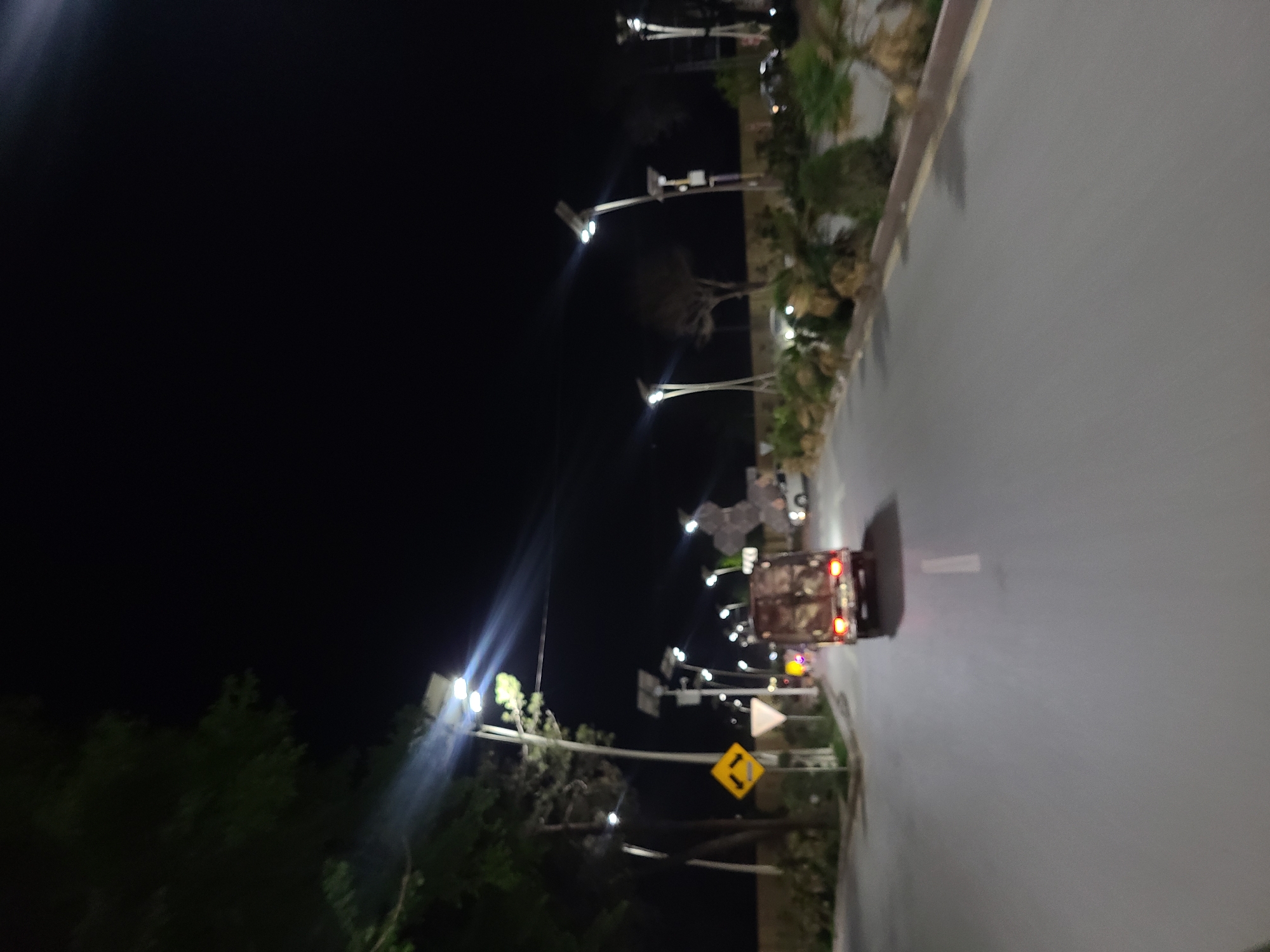 The image displays a blurry, nighttime outdoor scene along a paved road in Quetta City Tehsil, Pakistan. The perspective appears to be looking down the length of the road.
On the right side of the frame, a wide, dark grey paved road is visible, possibly damp or reflective, with a white line painted along its surface. A small, open-sided vehicle, resembling a rickshaw or utility vehicle, is positioned on the road, showing prominent red taillights.
Along the left edge and bordering the road on the right, there are multiple tall streetlights with bright, white LED-like lights, casting strong beams and lens flares into the dark night sky. The intense artificial lighting indicates the scene is captured at night or late evening. Dark silhouettes of trees and bushes line the road and extend into the background.
Visible among the vegetation are two road signs: a yellow, diamond-shaped sign featuring two black arrows pointing in opposite vertical directions, and a partially obscured white triangular sign with a red border, typically indicating a yield. The overall setting suggests a busy urban or semi-urban street at night. Due to the blurriness, no specific individuals are discernible, and no explicit text is visible other than the symbols on the road signs.