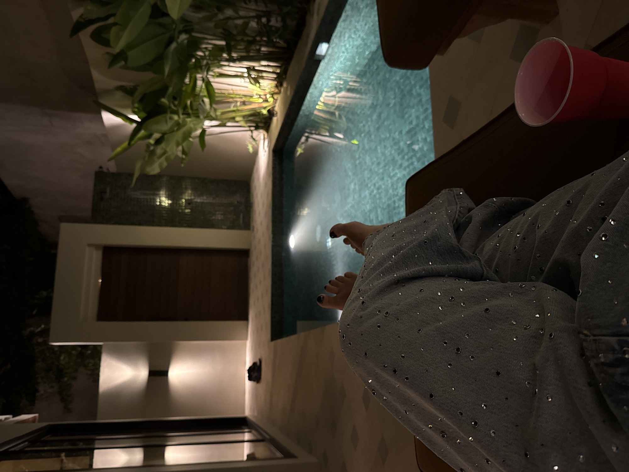 The image shows a scene at night, likely near a swimming pool in Kuta Selatan, Indonesia. A person is sitting with their legs extended towards the pool's edge. They are wearing dark blue denim pants with small reflective studs. Their toes, painted with dark nail polish, are visible near the water. A red plastic cup sits on a surface near the person. Lush green foliage and plants border the pool's edge. The architecture consists of white walls and accents, with a dark wood door visible in the background, illuminated by nearby lights. The swimming pool water reflects a bright light source, obscuring details within the pool itself.
