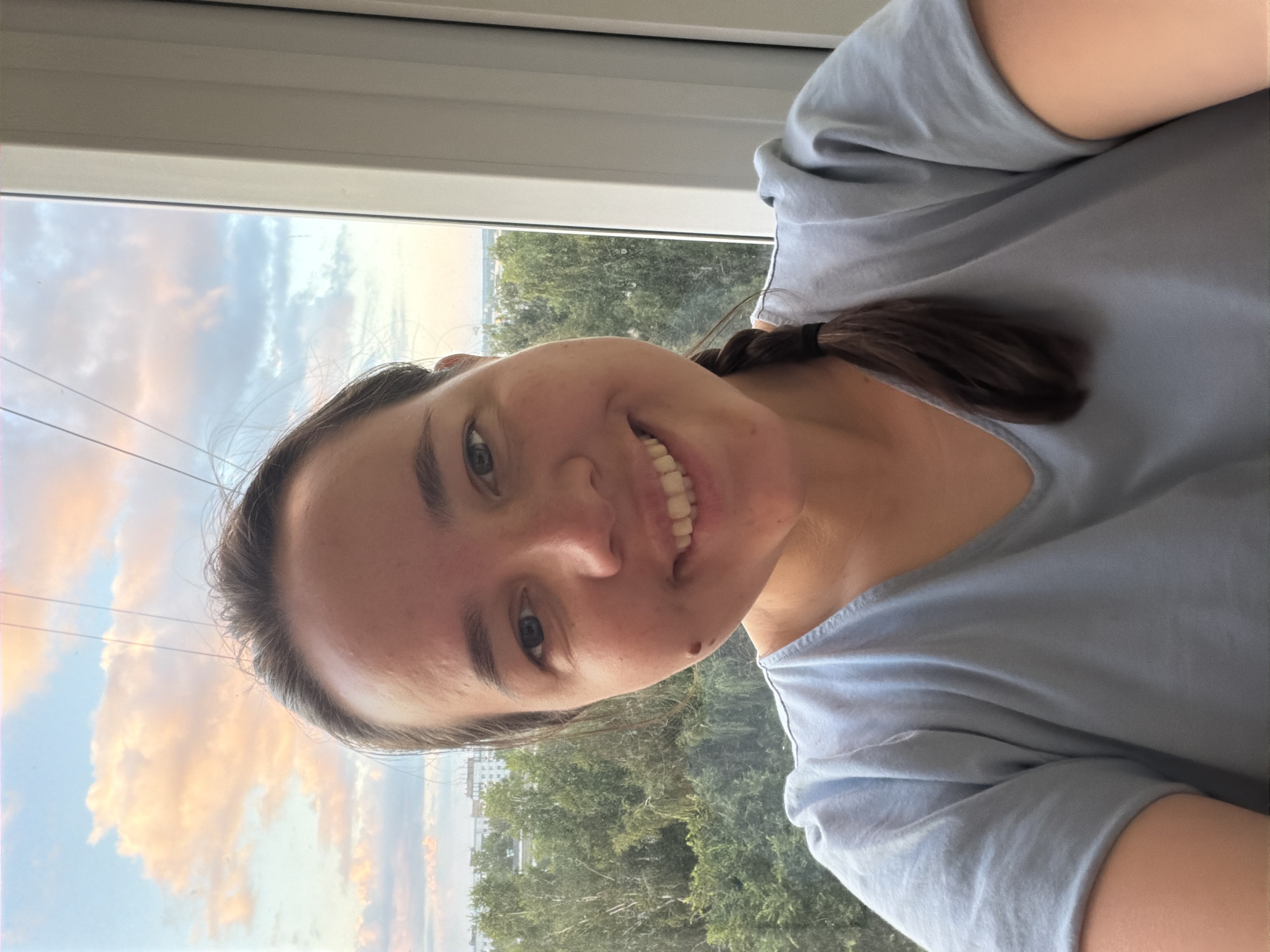 A woman is taking a selfie in front of a window, with trees and buildings visible in the background, suggesting an urban setting. The sky has clouds and appears to be either sunrise or sunset. The woman has brown hair, styled in a braid, and is wearing a light blue t-shirt. She is smiling directly at the camera. The location is specified as Chaikovsky, Russia.
