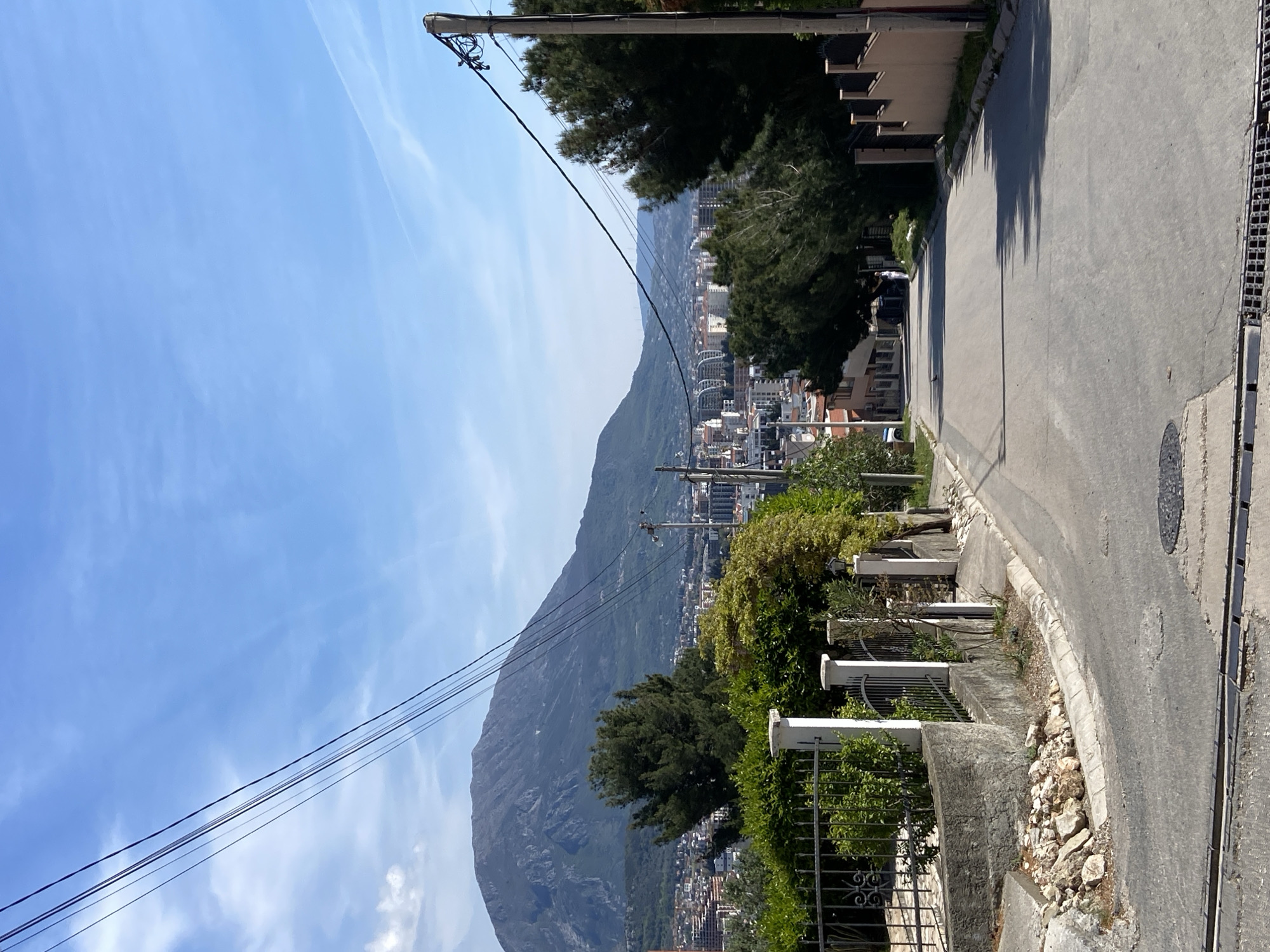 A medium, slightly angled shot depicts a residential street in Šušanj, Montenegro. The road, composed of gray asphalt, curves to the right and slopes downhill. The right edge of the road has a dark gray drainage grate, while the left edge has a low concrete barrier with patches of vegetation. Beyond the barrier, a mixture of trees, buildings, and the gray slopes of a mountain are visible. On the right side of the road is a light tan two-story building, partially obscured by trees. Power lines extend diagonally across the upper left quadrant, connected to a utility pole. The sky is clear blue with wisps of cirrus clouds in the upper left.
