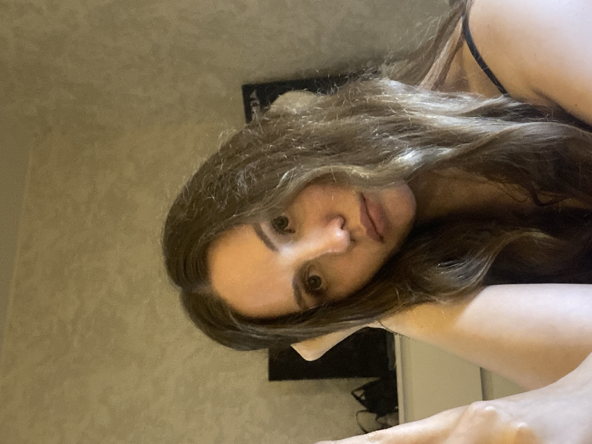 A woman with long, wavy brown hair is facing the camera. She has fair skin, brown eyes, and is wearing a thin strap. The background shows a beige wall and a dark, rectangular object. Based on the image context alone, no information is available to determine the location in the city of Izhevsk, Russia.