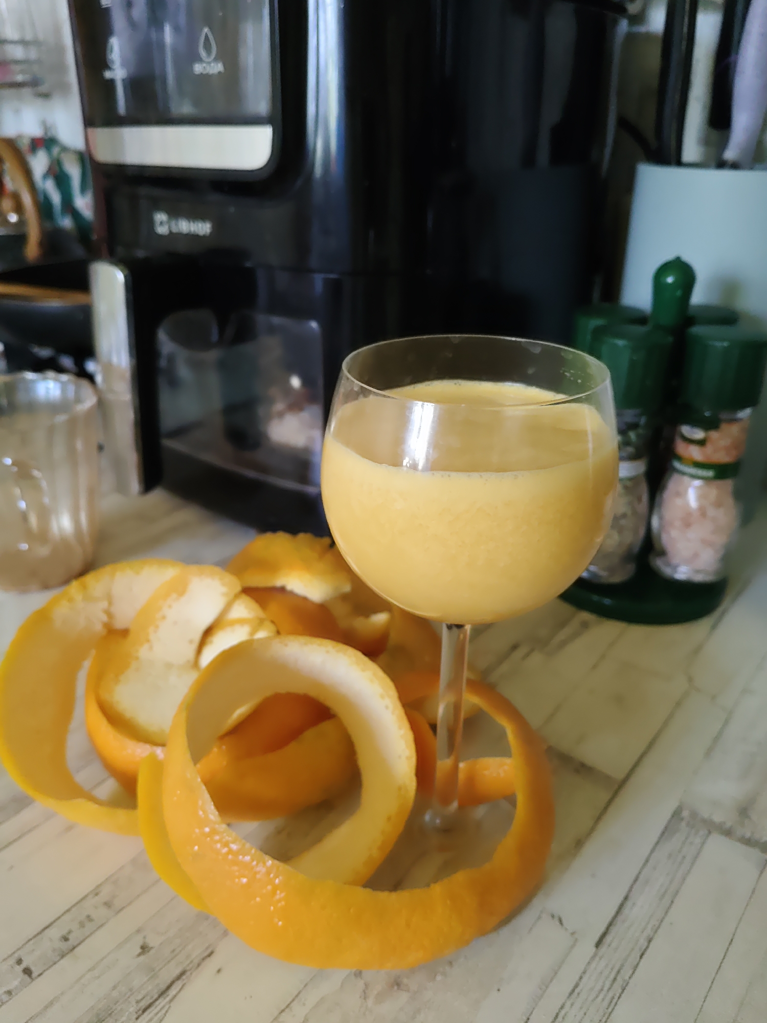 An indoor, close-up, eye-level shot shows a glass of orange juice on a wooden table. The glass is partially filled with the orange liquid and is on a stemmed wine glass. Around the base of the glass are several strips of orange peel, curled and uncurled, some with white pith still attached. In the background, a black coffee maker is visible, with a glass carafe and water reservoir. To the right of the coffee maker, a green rack holds two spice grinders. The overall environment appears to be a kitchen or dining area. The location is specified as Rostov-on-Don, Russia.
