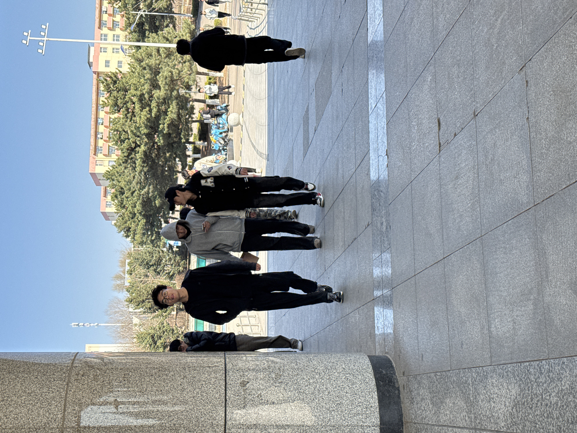 The image shows five people standing on a tiled sidewalk in Zhongtun, China. The people are wearing various clothing and facing away from the camera. In the background, there is a light pole, a building, and trees.