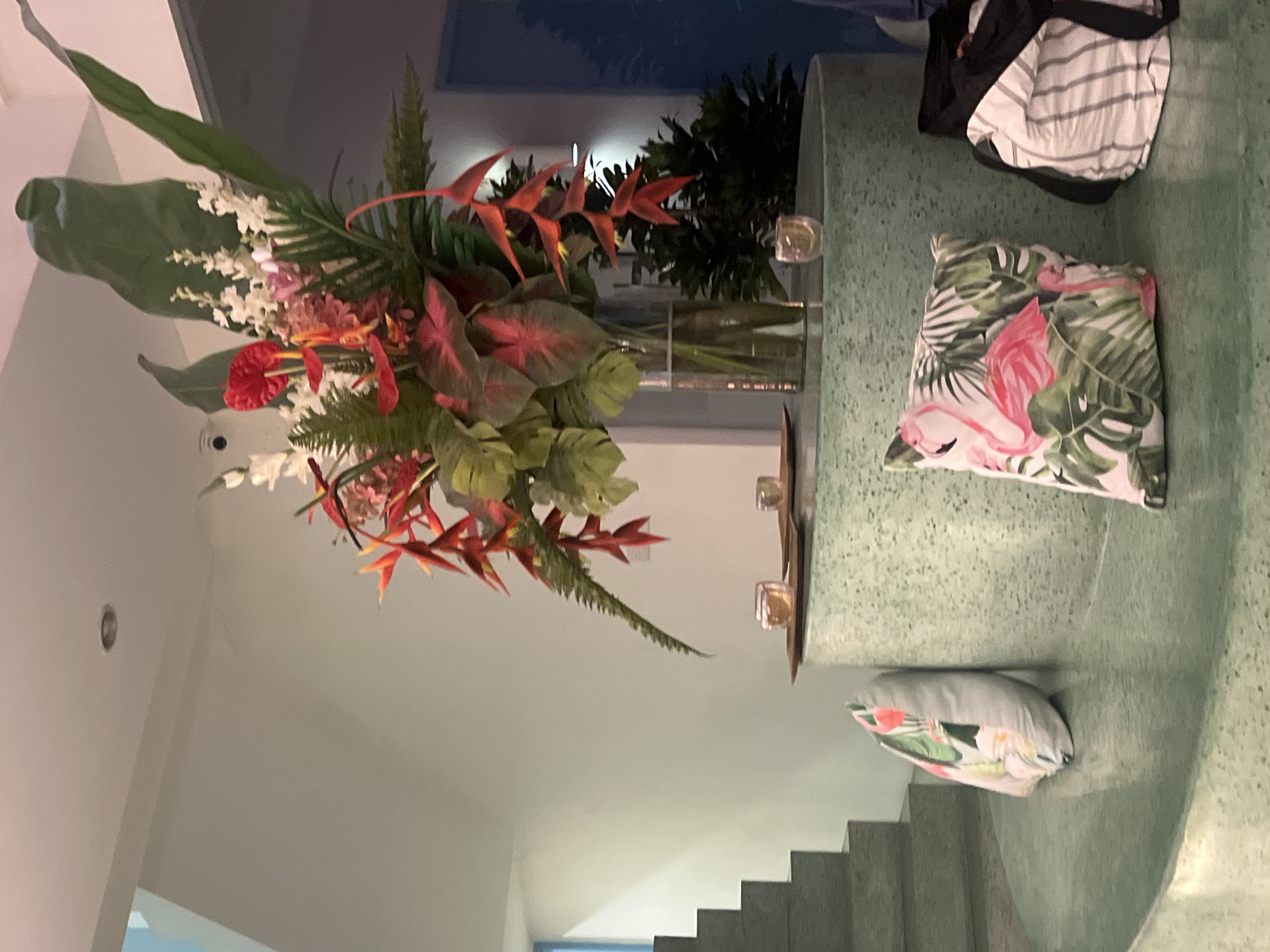 The image shows an interior setting, potentially a lobby or a staircase area, situated in North Kuta, Indonesia. A large floral arrangement dominates the scene, featuring an assortment of tropical flowers and greenery in a tall glass vase. A circular table with a speckled green surface is present, holding the vase. On the table are several small decorative objects resembling tealight holders. A striped bag sits near the edge of the table, and two decorative pillows with tropical patterns rest on the table's surface. The stairs lead up to the right and are also finished with a speckled green surface. The walls are painted a light color, and an inset light fixture is visible on the wall. The overall aesthetic is tropical and modern.