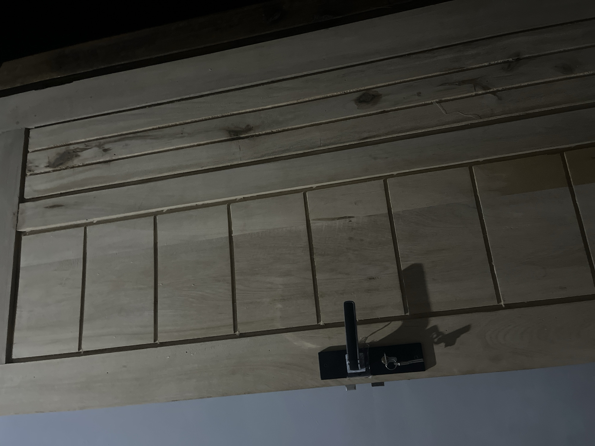 The image shows a close-up, angled view of a wooden door with horizontal paneling on the left side and vertical planks on the right. A black door handle with a silver mechanism is attached to the left side of the door, and a key is inserted into a lock below it. The door appears to be in a dimly lit interior, with a shadow of the handle and key cast on the door's surface. The background on the left is a plain, light-colored wall, while the right side of the image fades into darkness, suggesting the presence of a dark room or hallway. The overall impression is of a simple, functional doorway, possibly in a residential or commercial building. No specific landmarks or context are visible to place this image in Kebumen, Indonesia, or any other specific location.