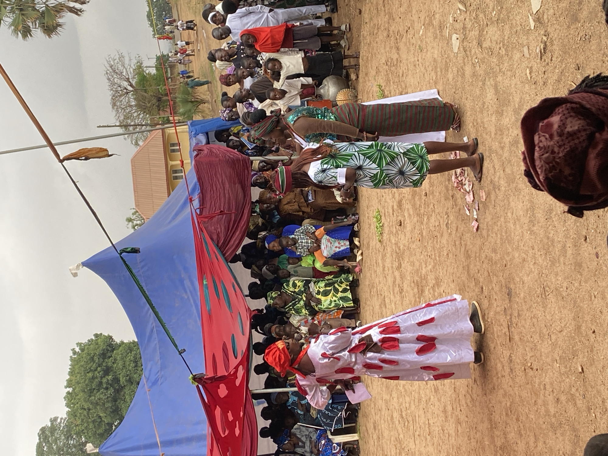 The image shows an outdoor event in Riyom, Nigeria. A large crowd of people is seated under a blue and red tent. Two women stand on the ground in front of the crowd. One woman wears a white dress with red polka dots and a red headscarf. The other woman wears a green and white patterned dress. Currency lays scattered on the ground near them. In the background, more people stand watching, and a building is visible. The ground is dirt.