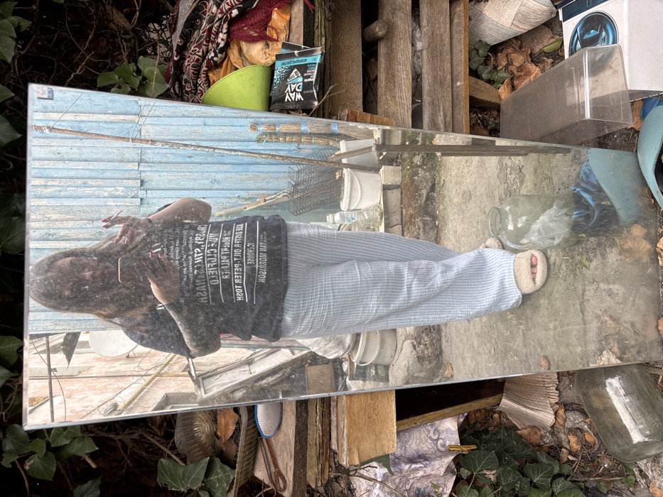 A large, rectangular mirror stands upright in a cluttered outdoor area in Chornomorskoe, Ukraine, reflecting a full-length image of a person. The reflected individual is a female with dark hair, wearing a dark t-shirt with white text (reversed), light blue and white striped pants, and light-colored fuzzy slippers. Tattoos are visible on both forearms. She holds a black smartphone horizontally in her right hand and a lit cigarette in her left.
The reflected background features a weathered light blue corrugated metal fence or wall, a lighter-colored building wall to the left, and a visible satellite dish. Various objects are reflected, including a long wooden pole, a metal grilling rack, plastic buckets, and bottles. The ground in the reflection is a mix of concrete and dirt.
Outside the mirror's frame, to the left, dense green ivy covers part of a wall or ground, alongside stacked wooden planks, a metal bowl, and a blue-handled brush. A large glass jar lies on its side at the bottom left. To the right of the mirror, a heavily cluttered area displays piled textiles, including rugs or blankets, a bright green plastic bowl, various plastic bags (one black bag visible with "YAW DAY" text), and books or magazines, all resting on a wooden structure. The ground around the mirror is composed of dirt, fallen leaves, and concrete, with several large glass jars and broken plastic items scattered.
