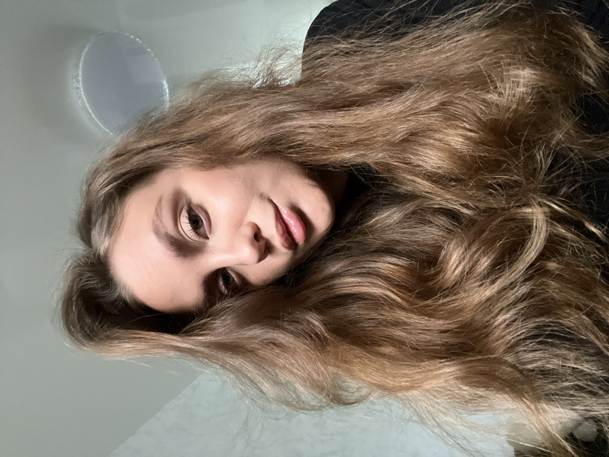 A young woman with long, wavy, light-brown hair is looking at the camera from a low angle. Her head is turned slightly to her left. She has fair skin, brown eyes, and full lips. She is wearing a dark-colored top. The background appears to be a light-colored ceiling with a round light fixture. The location is undetermined, and there is no evidence to suggest it is Chaikovsky, Russia.