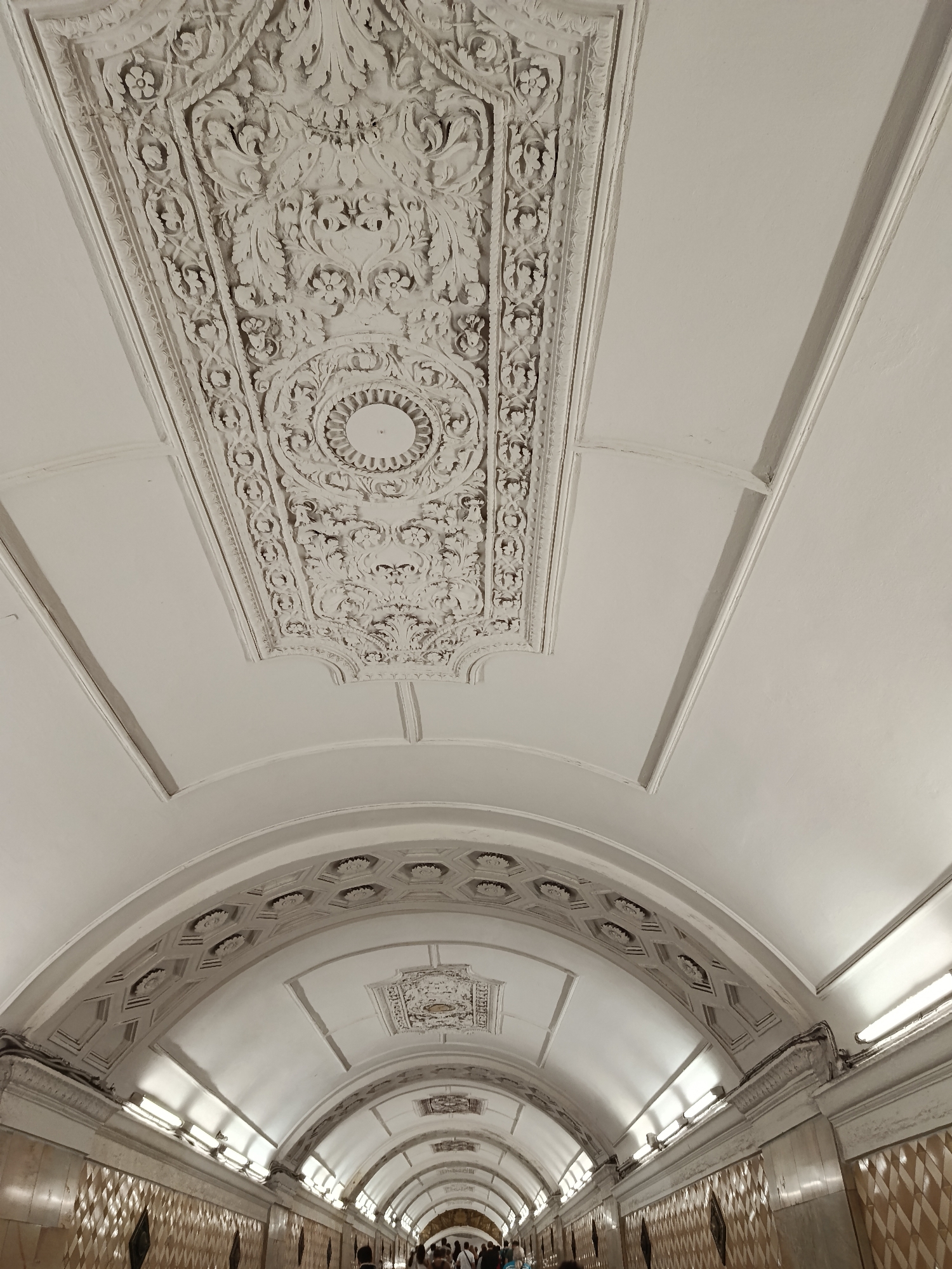 The image displays a low-angle perspective of an ornately decorated underground passageway or subway station, looking upwards and along its length. The ceiling is the primary subject, featuring intricate architectural details.
In the foreground and upper center, a large, rectangular panel of highly detailed white plasterwork or stucco is prominent. This panel is adorned with dense floral, leaf, and scroll motifs, bordering a central circular element that appears to be a decorative light fixture or medallion. Surrounding this central panel are smooth white ceiling sections with simple linear moldings.
Further along the passageway, the ceiling transitions into a series of arches. The curved surfaces of these arches are also decorated with repetitive geometric patterns, including hexagonal shapes containing central floral or starburst designs. Artificial lighting is provided by rows of fluorescent tube lights mounted along the upper edges of the side walls, illuminating the entire space.
Below the lighting fixtures, the walls are clad with patterned tiles, exhibiting a diamond or checkered texture. In the distant background, at the bottom of the image, several people are visible walking through the passageway, indicating it is an active public thoroughfare. The overall aesthetic suggests a historical or classical architectural style, characterized by its grandeur and detailed craftsmanship. No text is visible in the image. The indoor setting and artificial lighting provide no clues about the time of day.