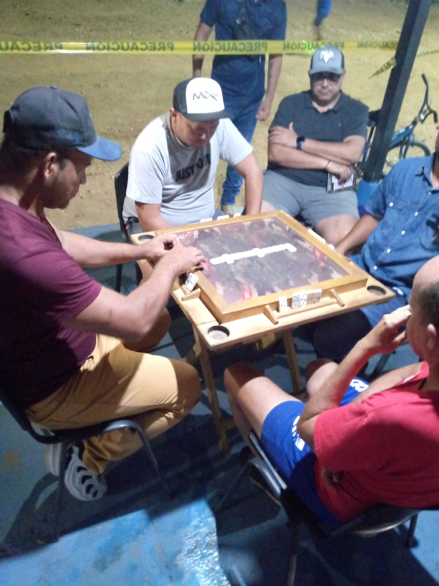 Four men are gathered around a folding wooden table playing dominoes outdoors. The game surface is clear and dominoes are visible. A fifth man stands behind them, and a sixth man sits and watches. Yellow caution tape is strung in the background. A bicycle is partially visible to the right. The location is Juan Griego, Venezuela, based on the caution tape.